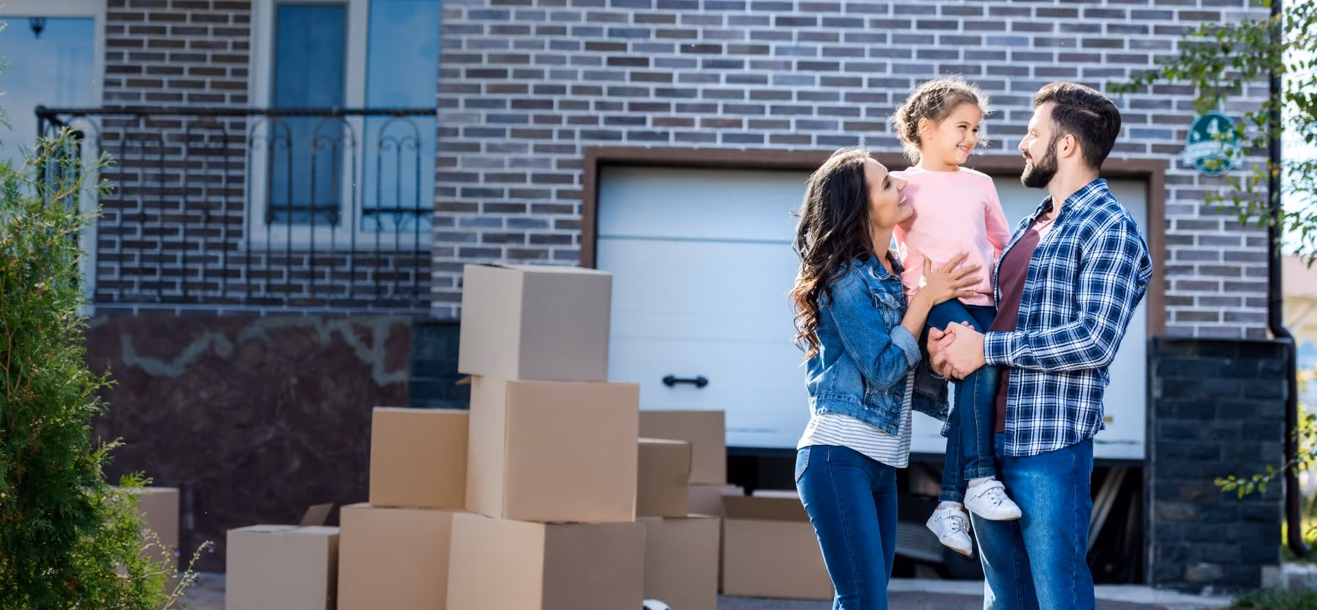 Smiling family of three with moving boxes outside a brick house with garage door.