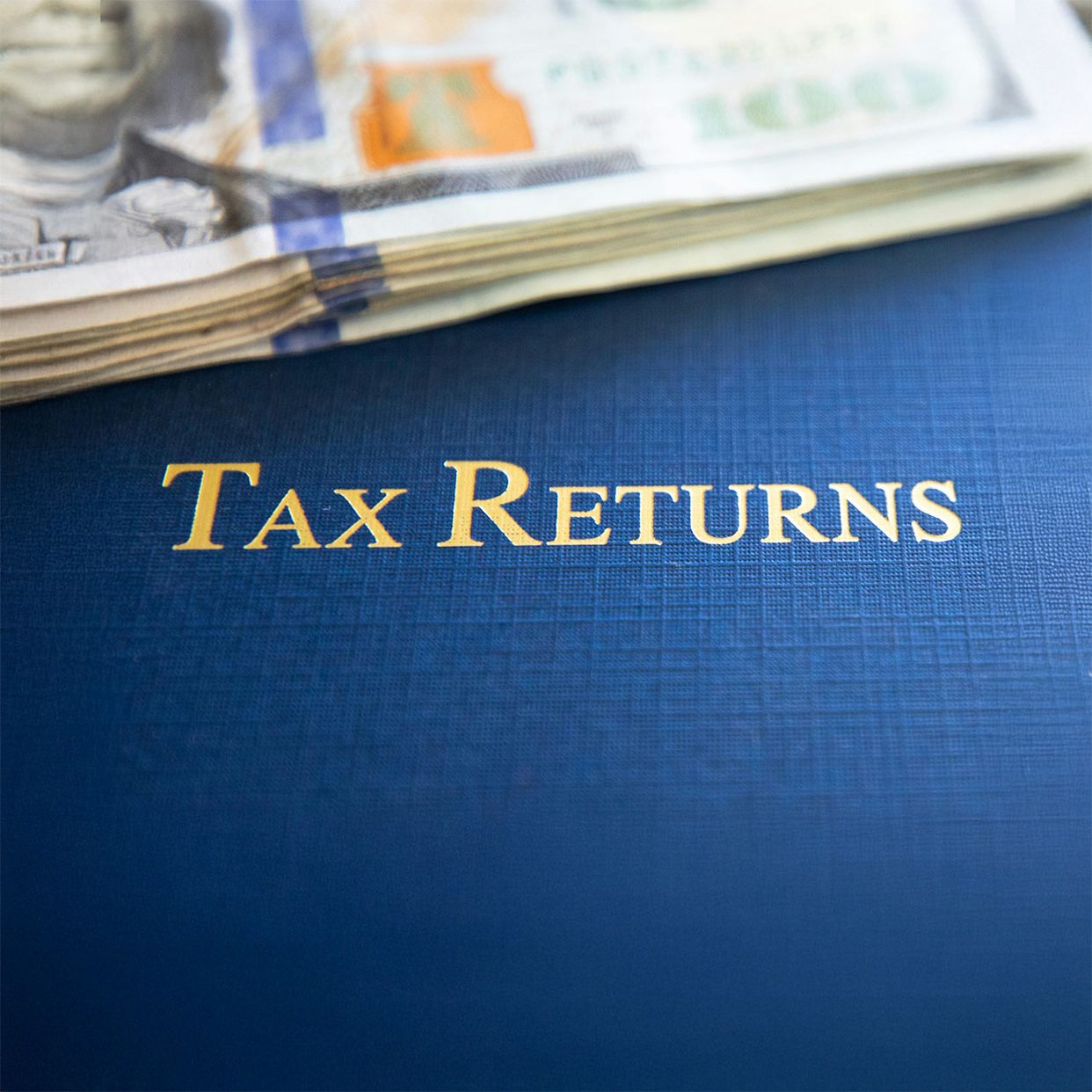 Blue folder labeled Tax Returns with a stack of hundred dollar bills, representing tax preparation and financial services.