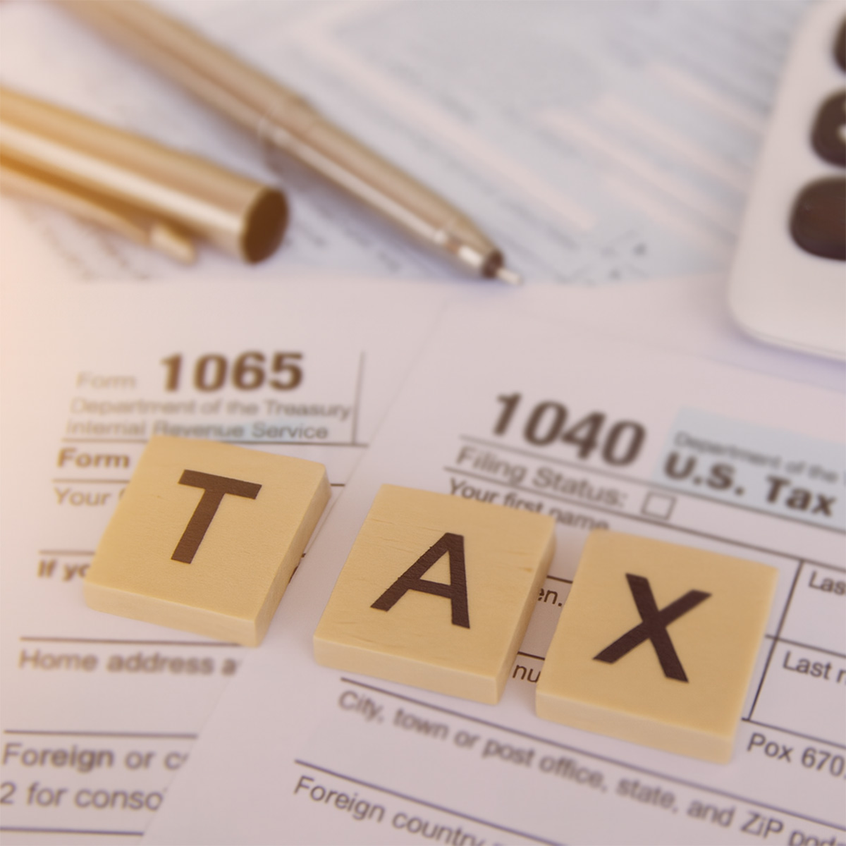 Wooden letter tiles spelling “TAX” resting on IRS Form 1040 and Form 1065, with a pen and calculator partially visible behind them.