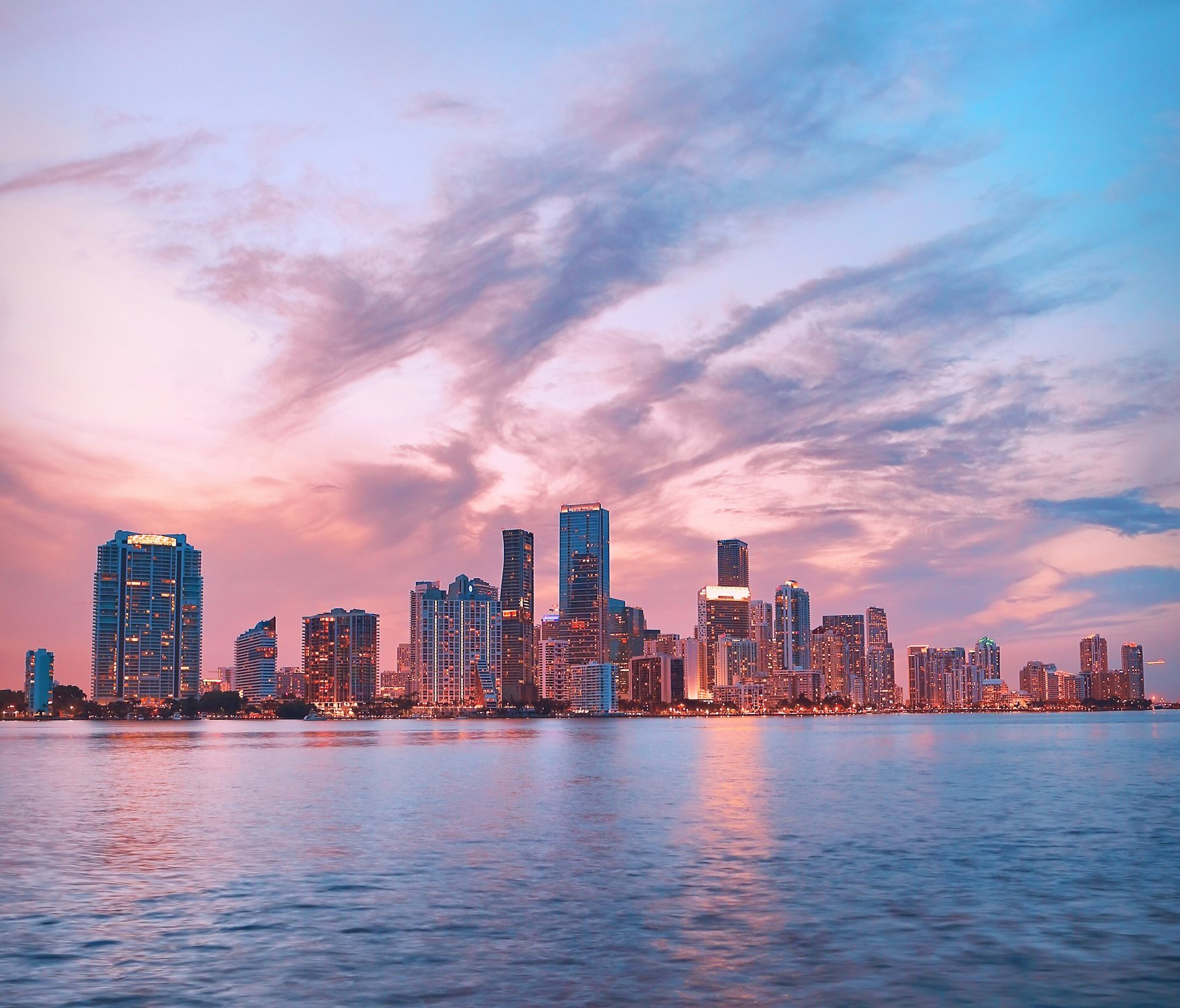 Miami skyline at sunset, with tall buildings reflecting warm pink and orange light across the water under a partly cloudy sky.