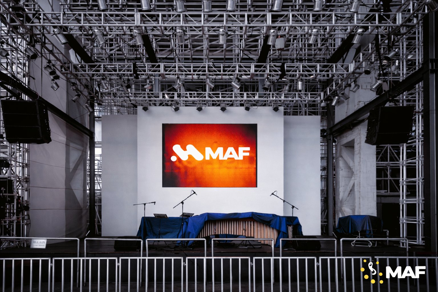 Scène de concert vide avec un grand écran MAF, des microphones et une structure de treillis métallique avec des lumières.
