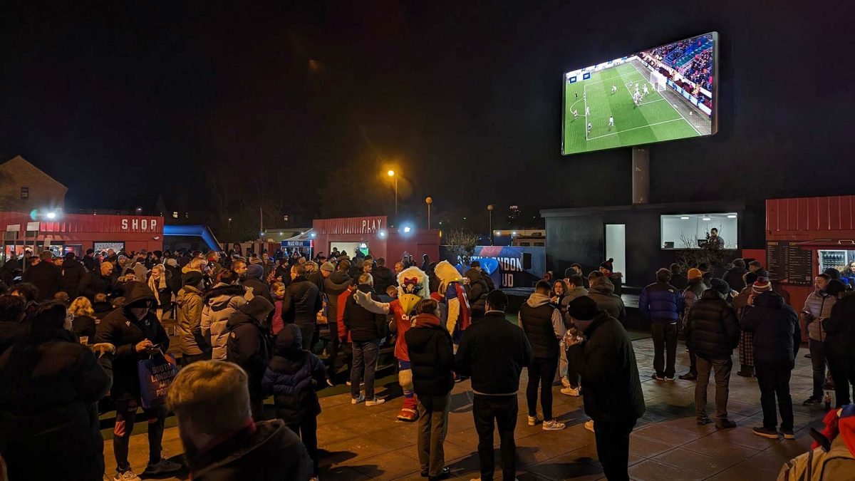 Grand écran LED extérieur diffusant un match de football, avec une foule de supporters et des stands de nourriture la nuit.
