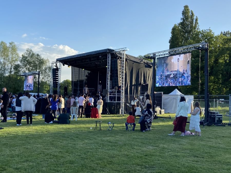 Une scène de concert extérieure avec deux grands écrans LED affichant des artistes, et des personnes regardant le spectacle sur l'herbe.