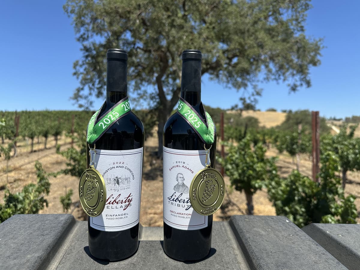 Two bottles of Liberty wines with 2025 gold medals hanging on green ribbons, displayed outdoors in a vineyard.