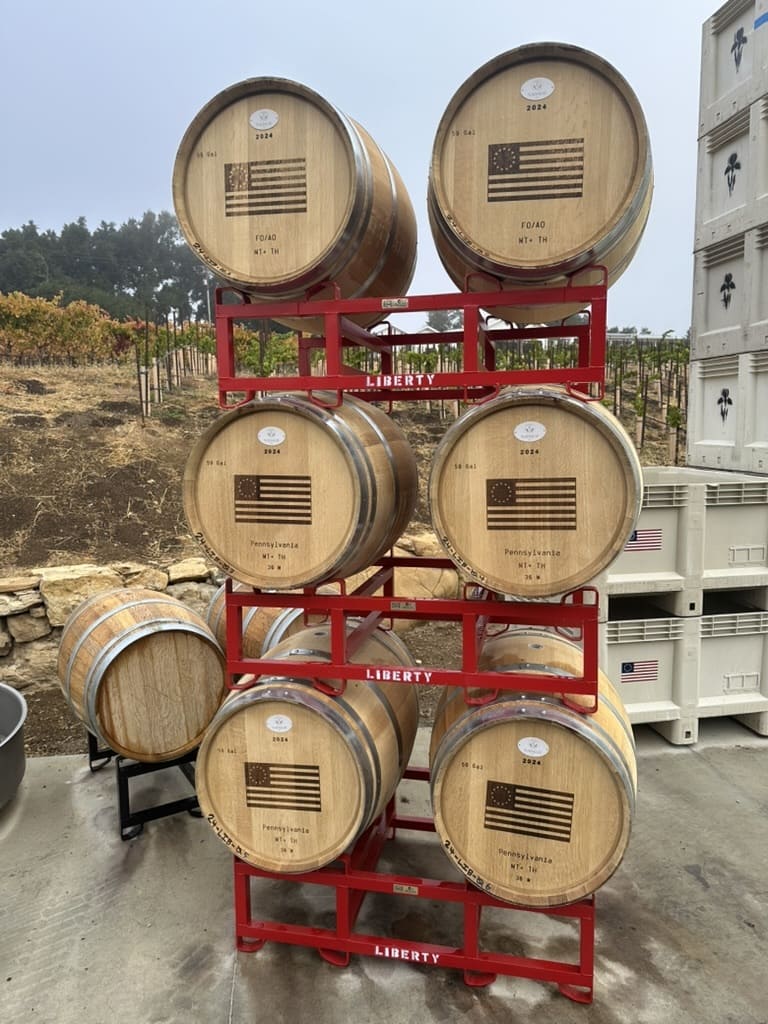 Six wooden barrels with an early American flag and text, stacked in red metal racks labeled Liberty, outdoors near vineyard rows.