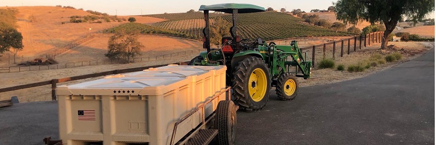 Green tractor towing large cream-colored crates on a country road beside vineyards under a setting sun.