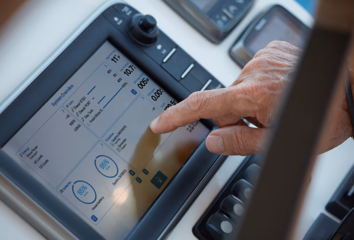 A close-up of a person’s hand interacting with a touchscreen control panel on a boat, displaying a system overview with energy, battery, and generator data.