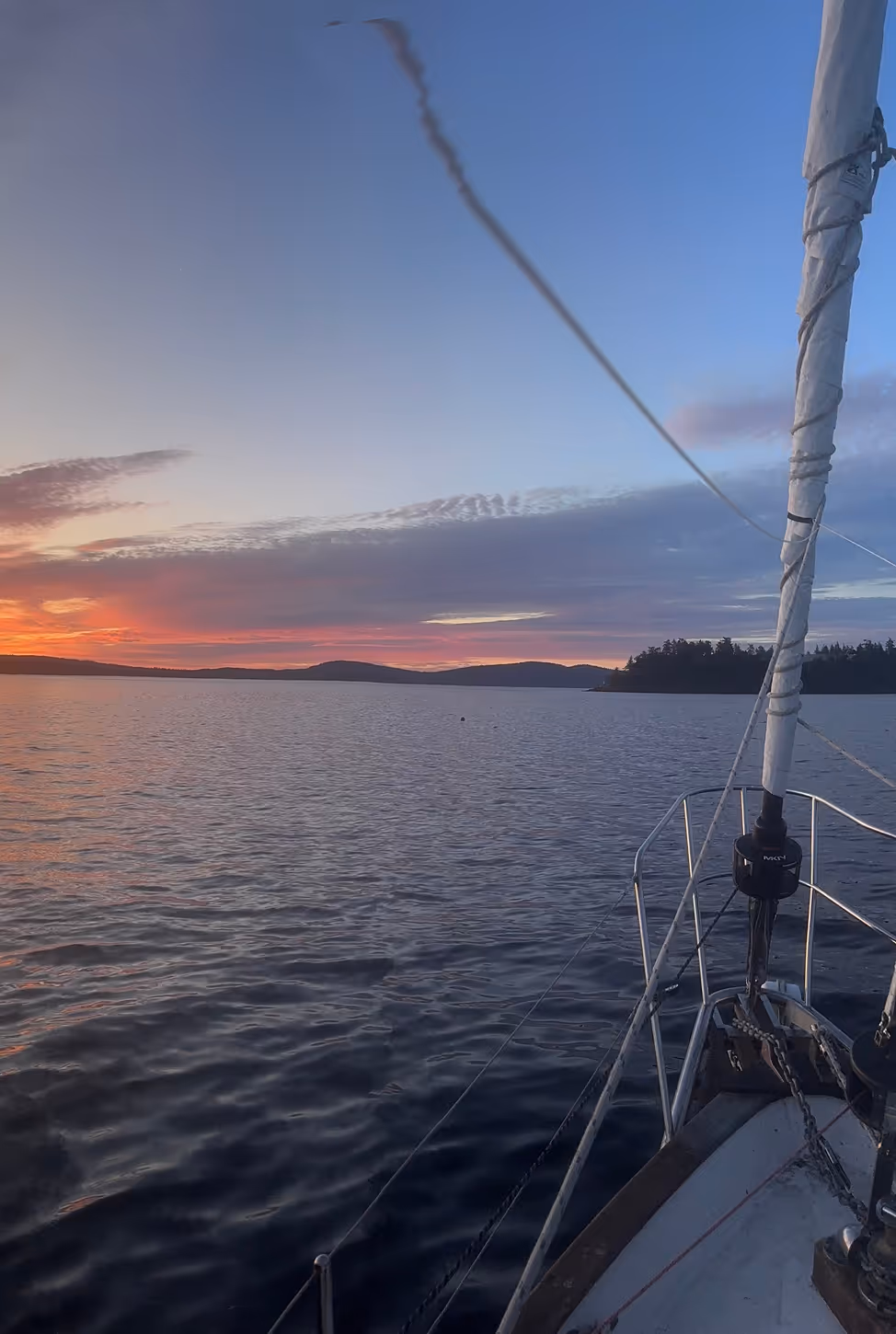 View from the bow of a sailboat at sunset, with colorful clouds over the horizon and calm water reflecting the fading light.