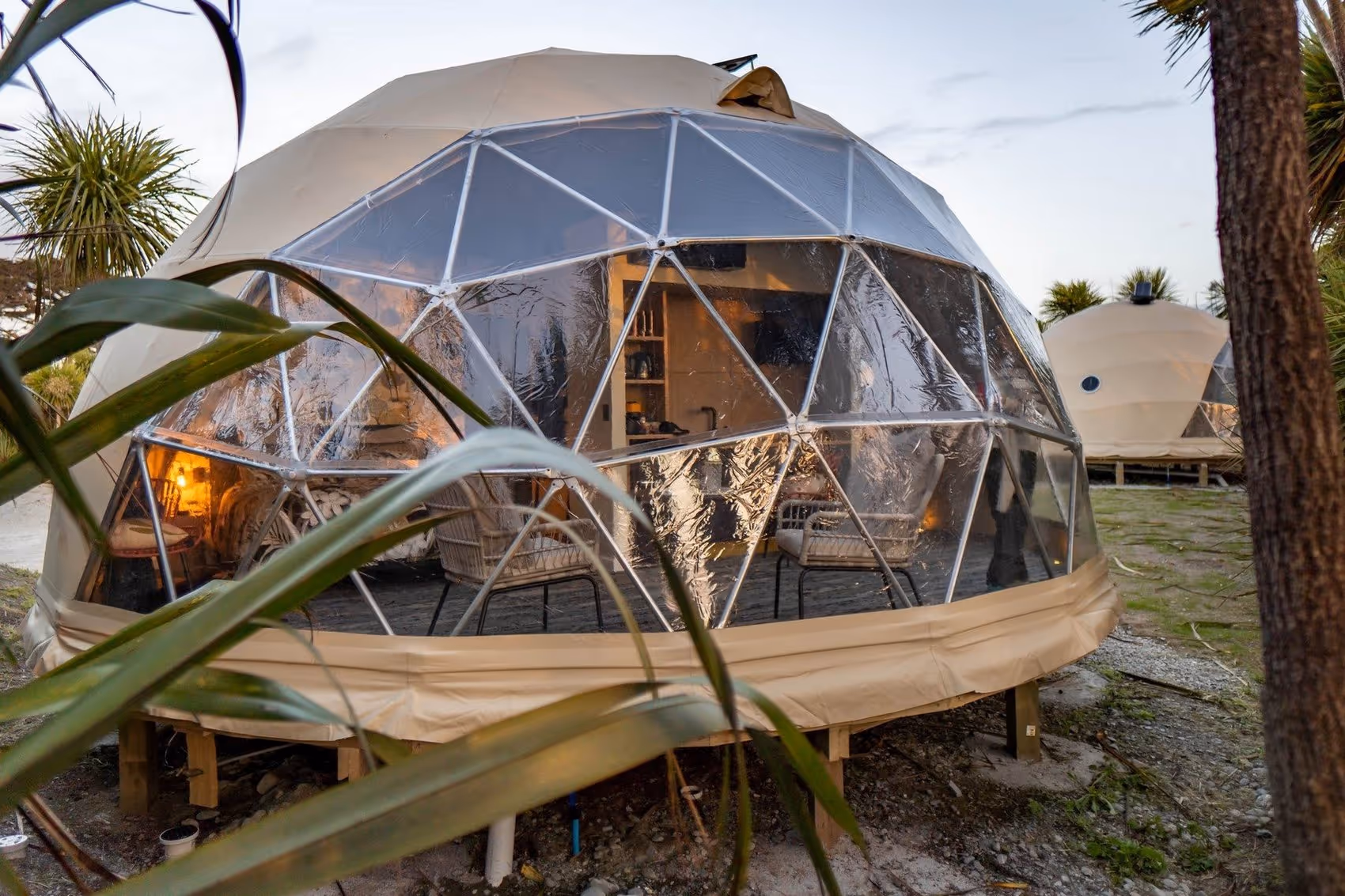 Transparent geodesic dome with warm internal lighting, surrounded by plants and trees. Another dome is visible in the background, creating a cozy, serene atmosphere.