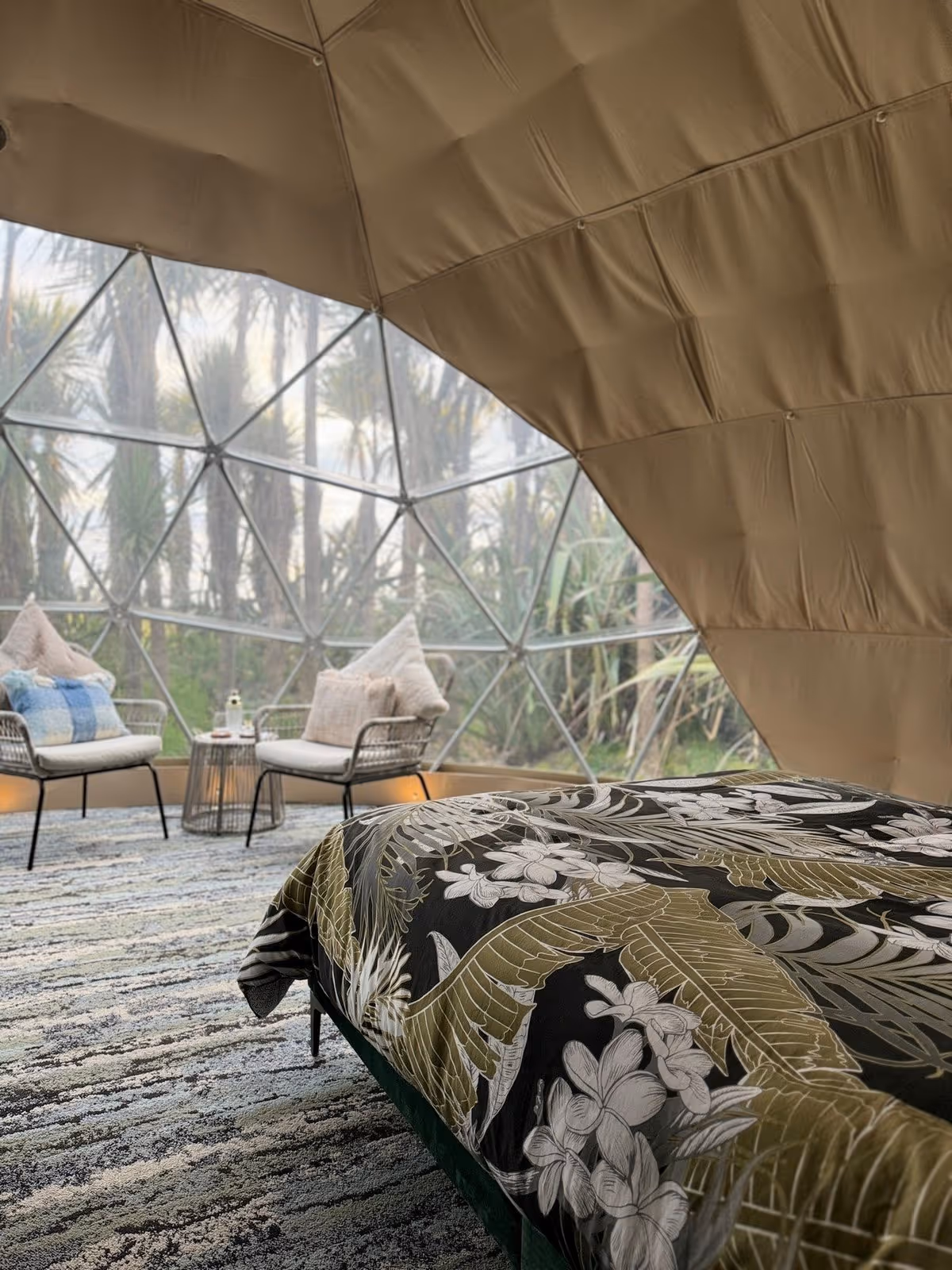 Cozy dome interior with a floral-patterned bed. Two cushioned chairs and a small table face a large geodesic window, offering lush greenery views outside.