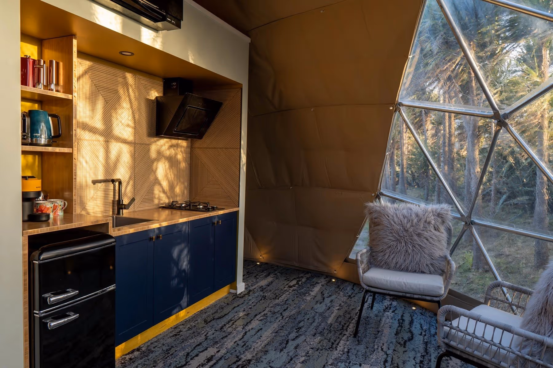 A cozy modern kitchenette inside a geodesic dome with a view of trees. Features include a blue cabinet, countertop, small fridge, chairs, and a furry cushion.