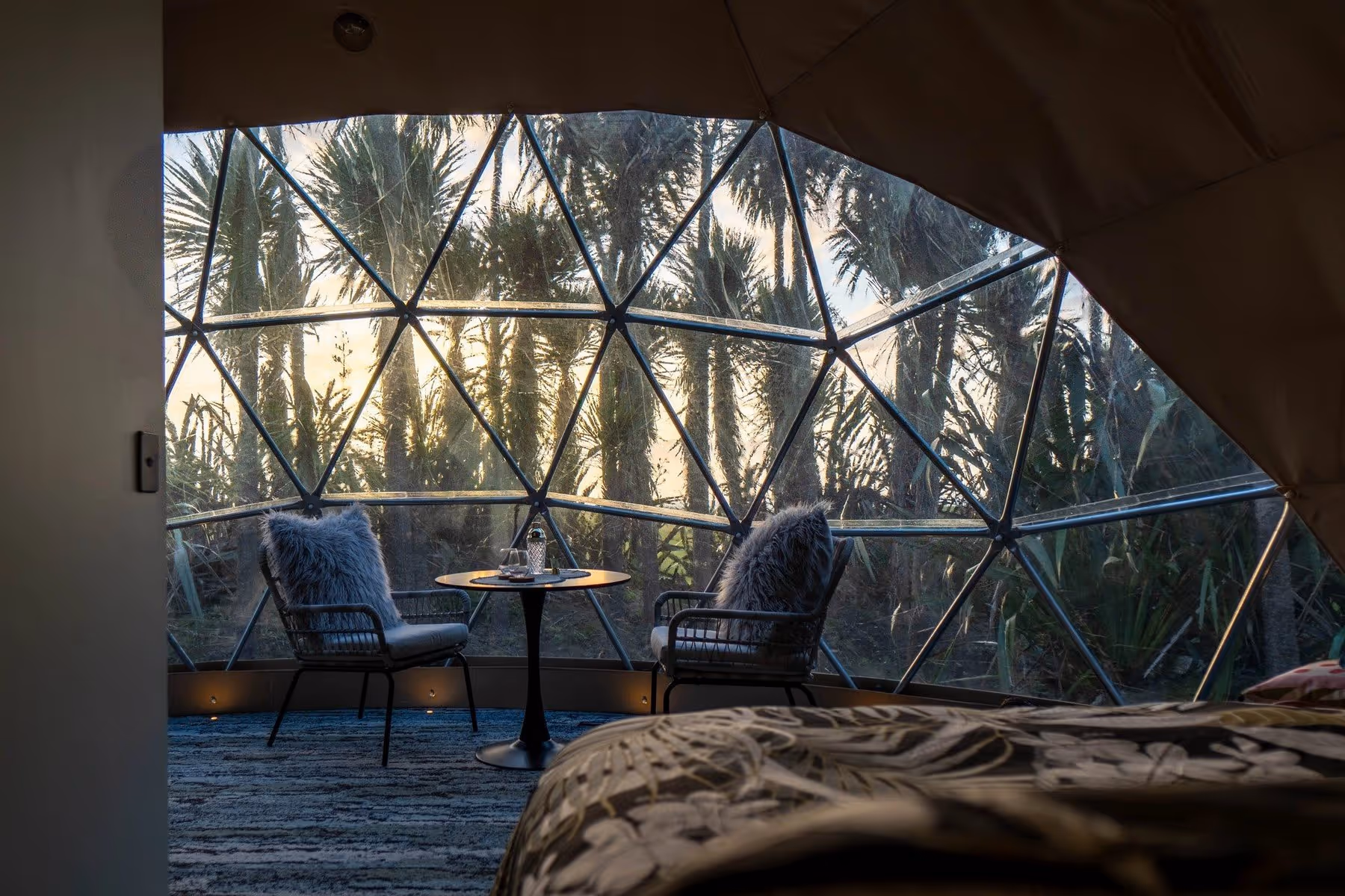 Cozy dome interior with a geodesic window showcasing lush palm trees at dawn. Two chairs and a small table create a serene, inviting atmosphere.