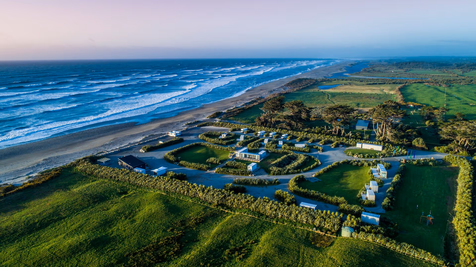 Aerial view of Ross Beach Top10 coastal retreat featuring blue cabins, lush greenery, and a sandy beach lined with ocean waves under a colorful sky.