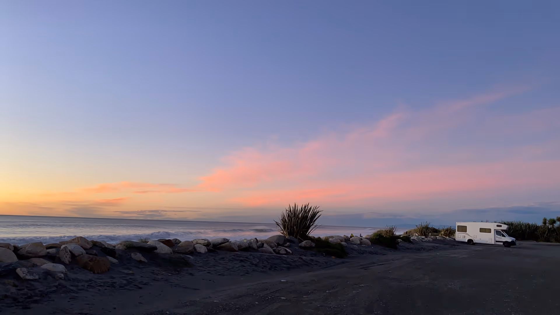 A tranquil beach scene at sunset, featuring a white camper van beside rocks and lush coastal plants, with soft pink clouds in the sky.