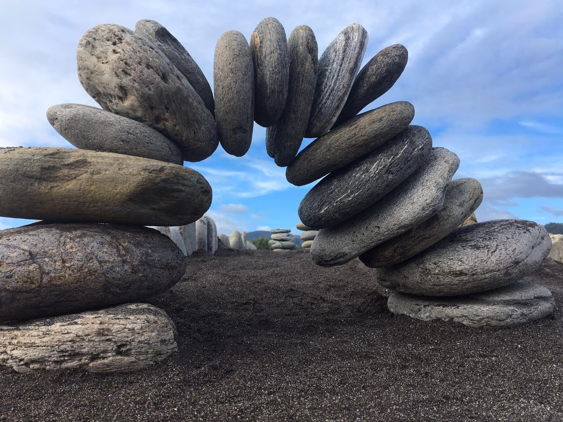 A carefully balanced arch made of round stones sits on black sand against a backdrop of cloudy blue sky.
