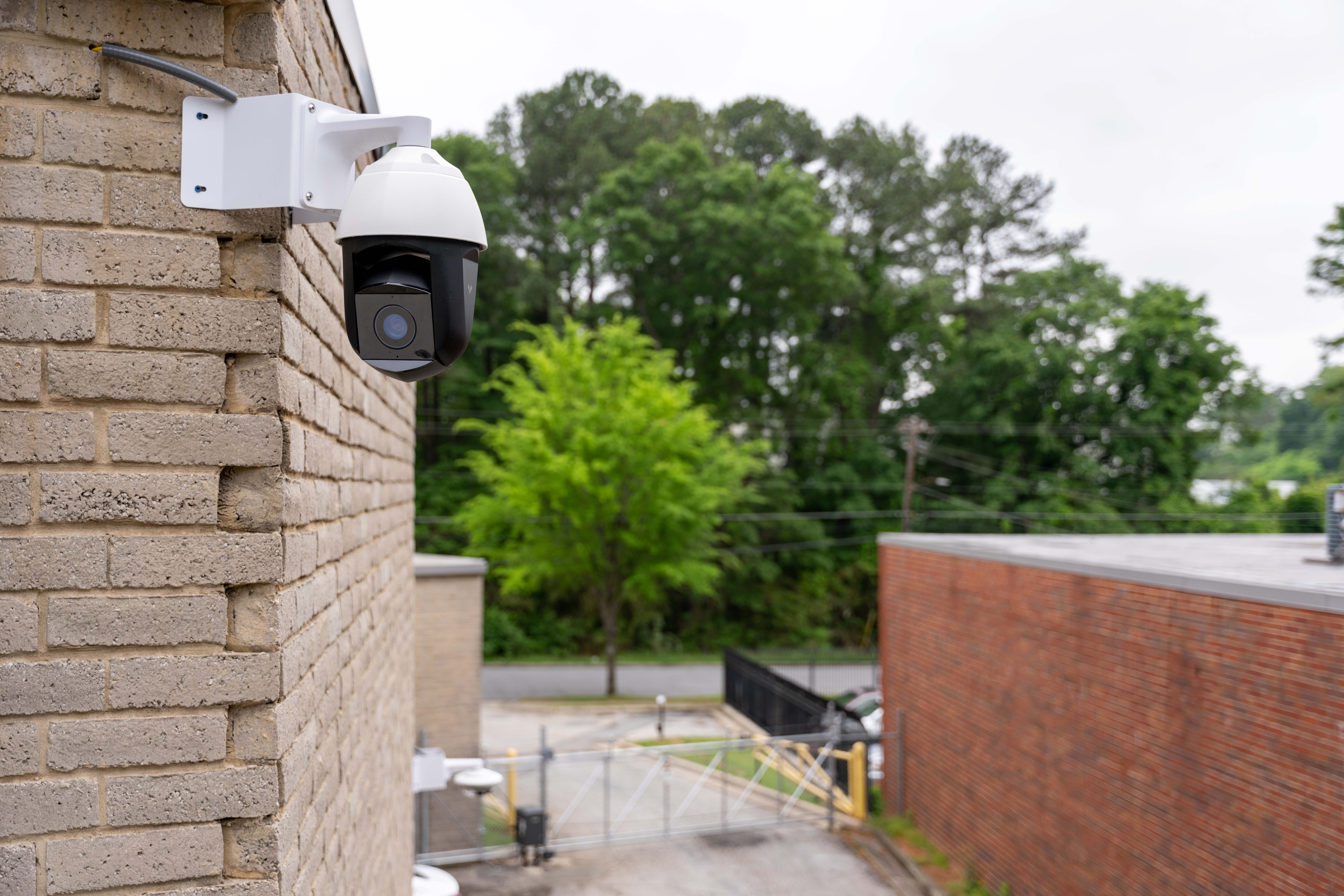 Surveillance camera mounted on a brick building overlooking a gated parking area with trees in the background.