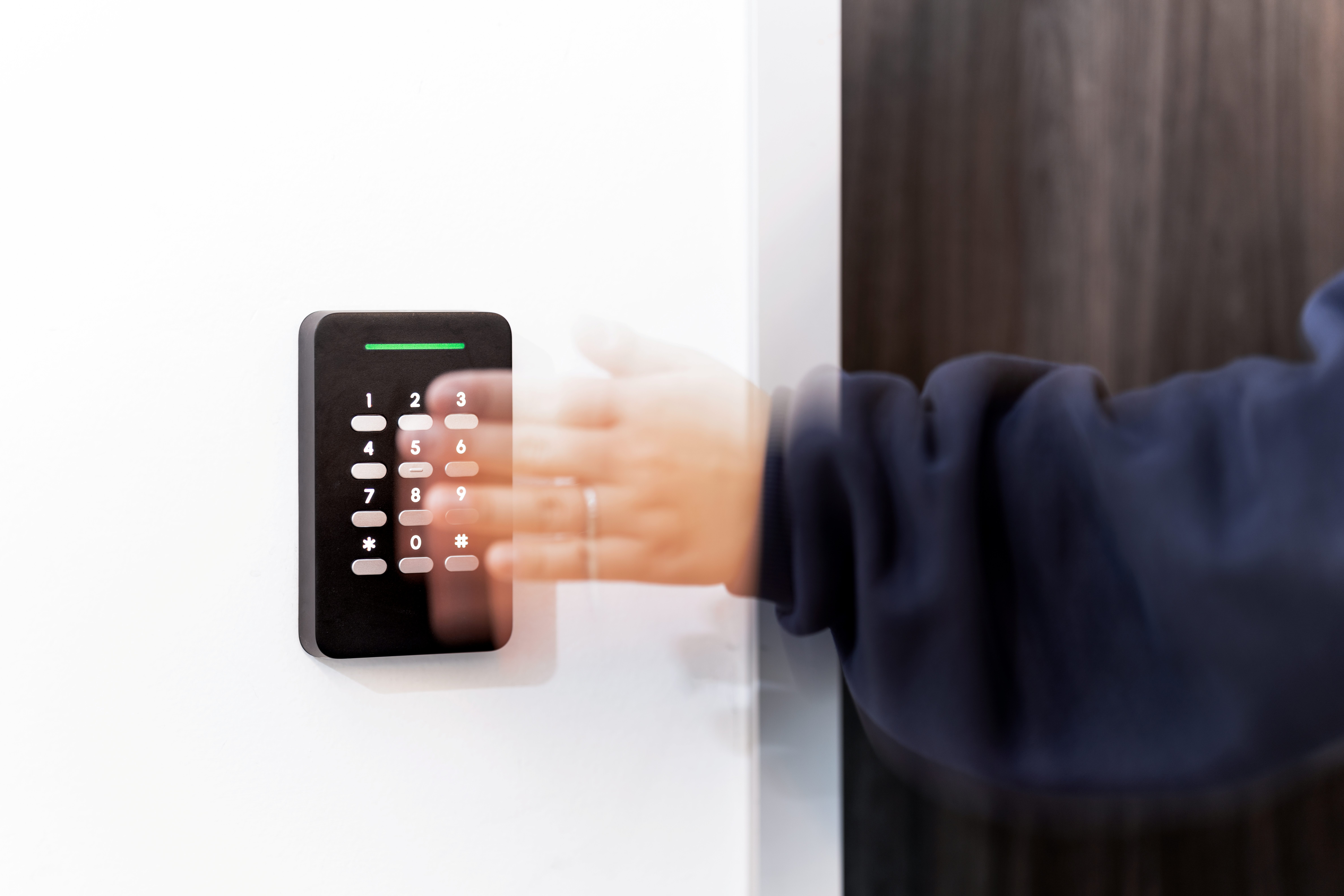 Person entering code on a black keypad access control mounted on a white wall.