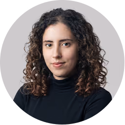 Woman with curly brown hair wearing a black turtleneck and neutral expression on gray background.