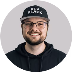 Smiling man wearing glasses, a black hoodie, and a black cap reading 'REV BLACK' on a gray background.