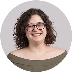 Smiling woman with curly brown hair, glasses, and olive off-the-shoulder top against gray background.
