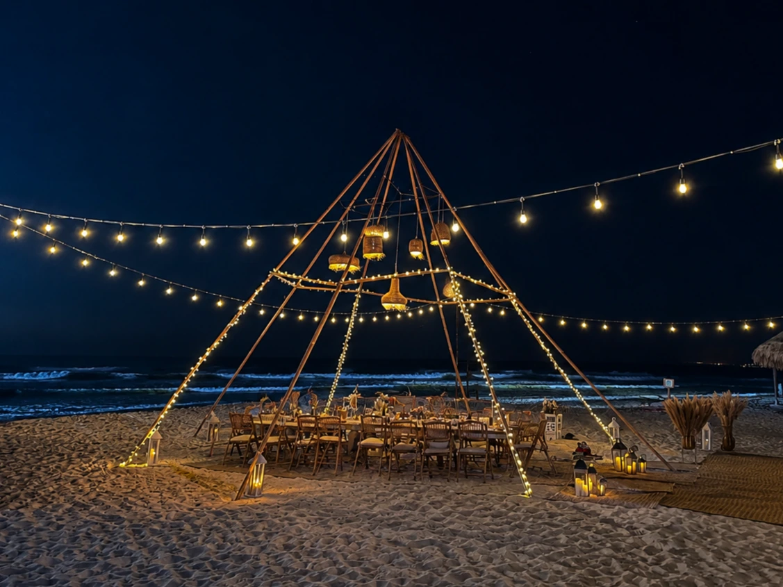 Completed beachfront dinner beneath a wooden teepee with woven lanterns, string lights and lounge seating.