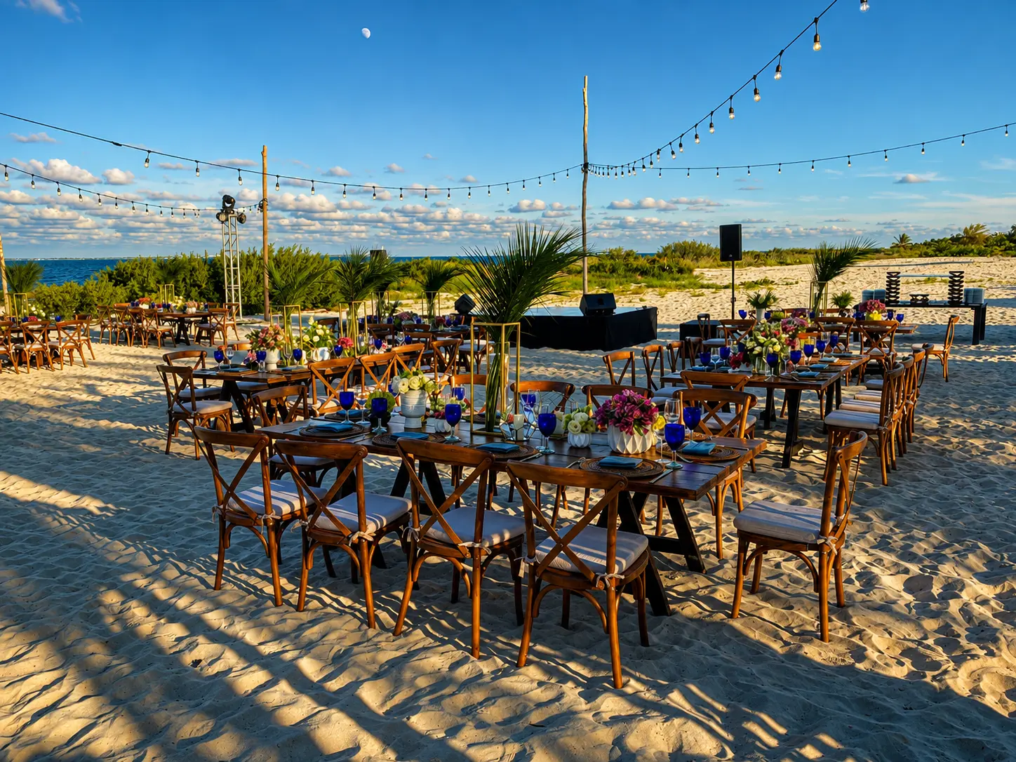 Beach tables are set with blue glassware, gold accents and tall floral arrangements matching the render.