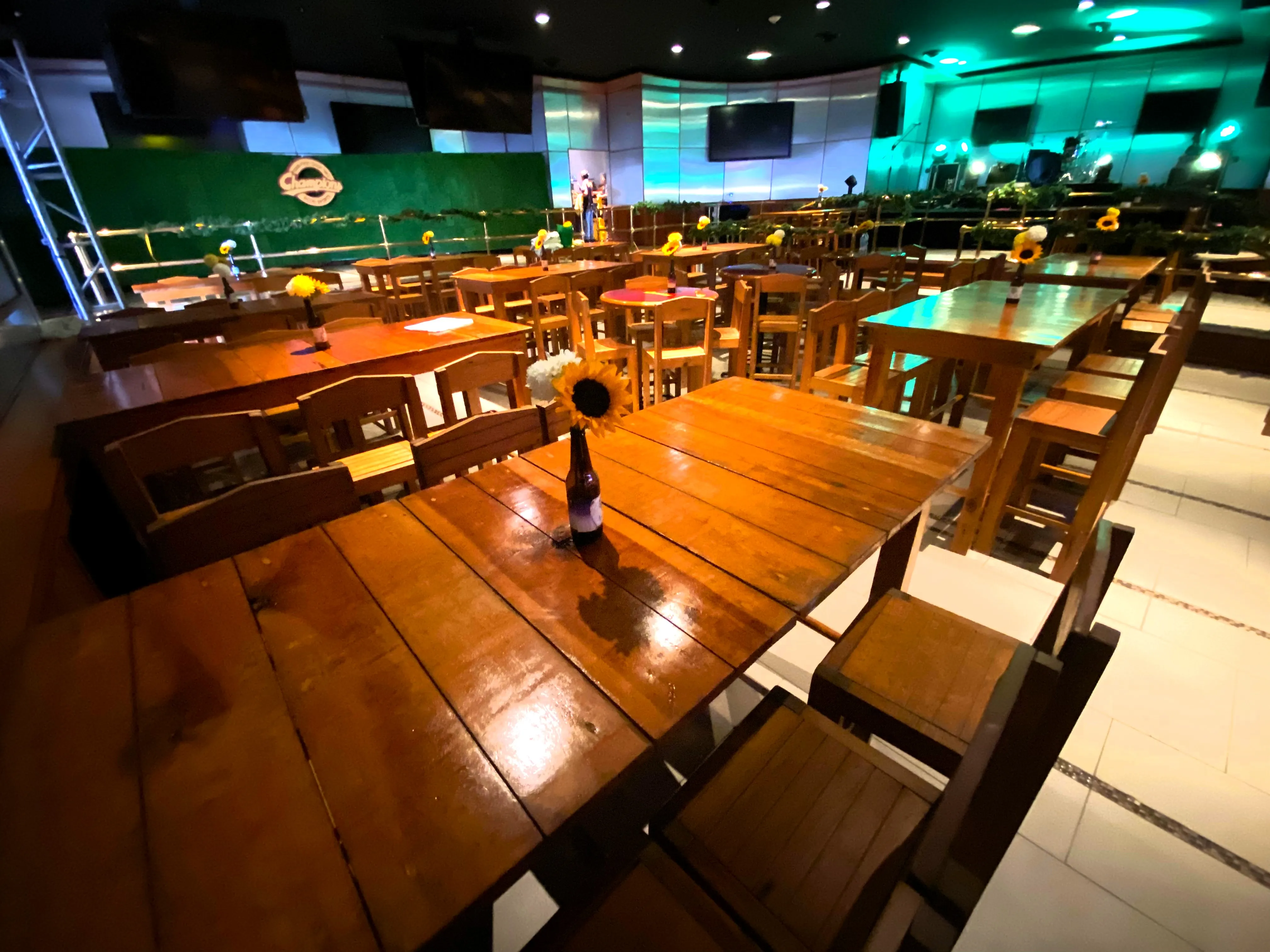 Wooden cocktail tables and stools fill an interior venue with a green wall and a stage in the background.