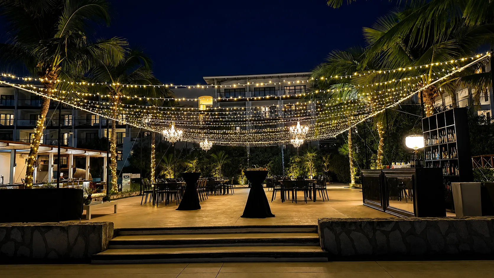 String lights and chandeliers stretch across an outdoor dining area with black tables set for a formal dinner at dusk.