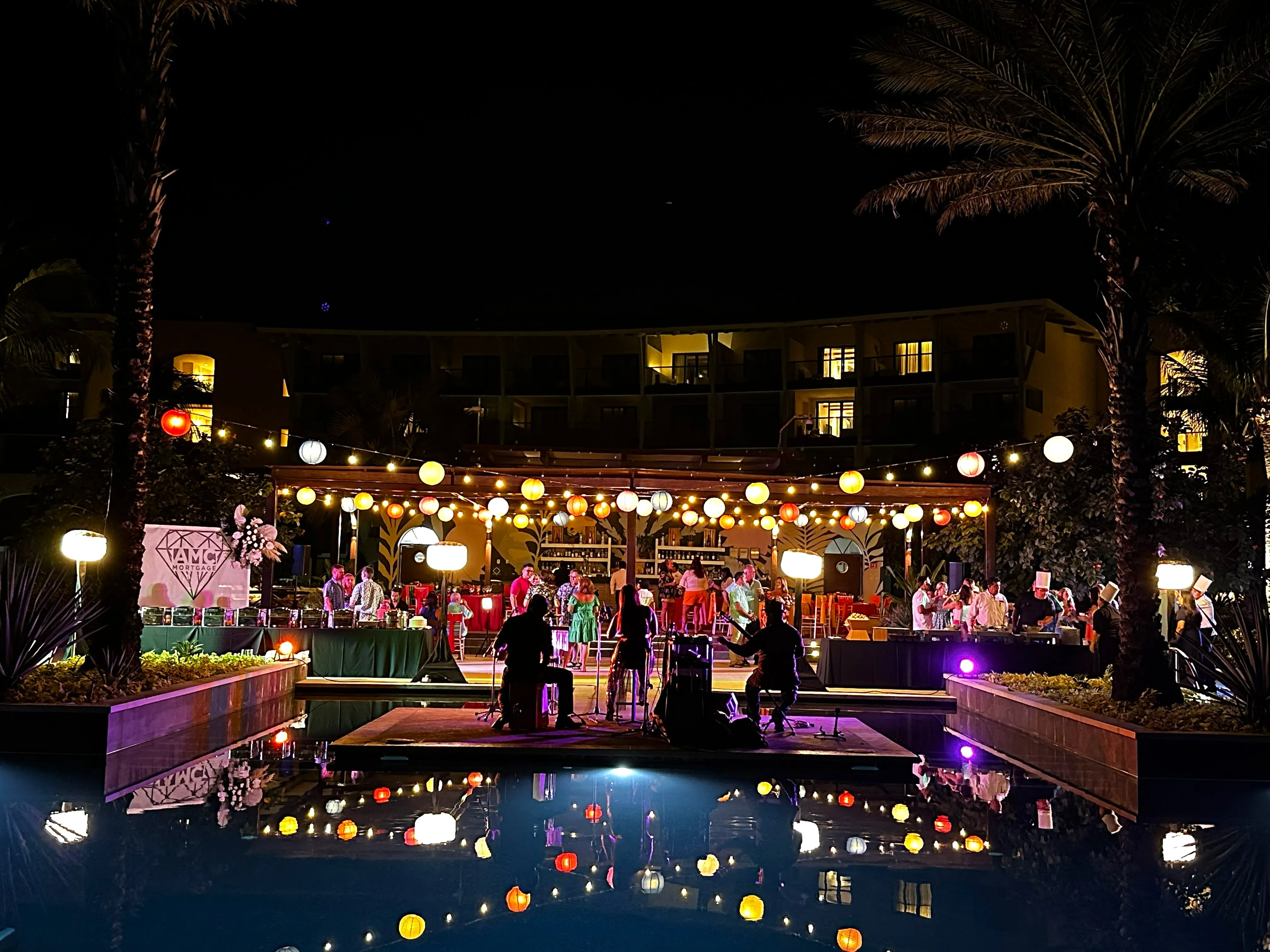 Colorful paper lanterns hang above a pool where musicians perform on a lit stage at night.