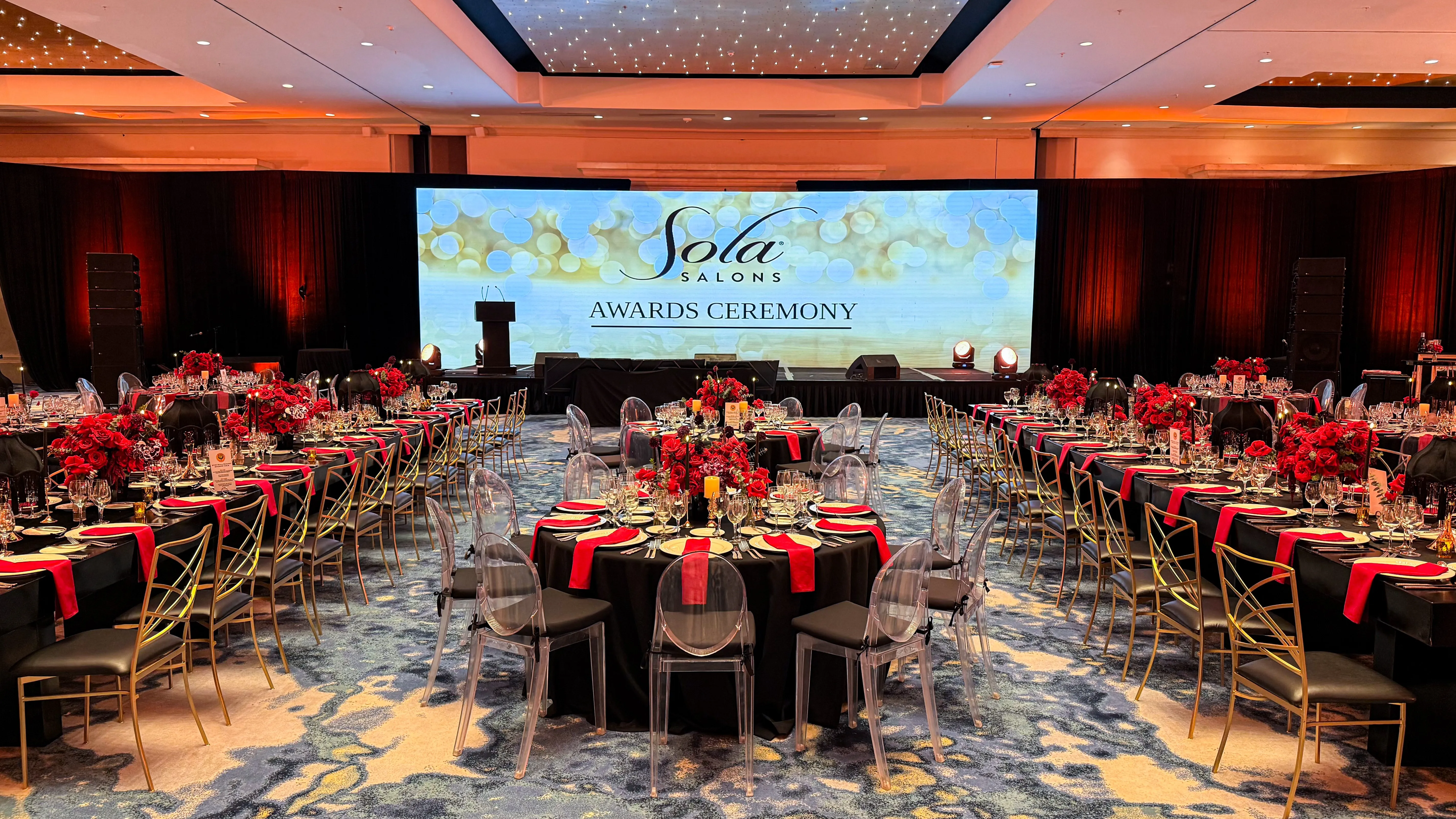 Long tables dressed in black with red rose centerpieces and gold chairs sit before an illuminated stage screen.