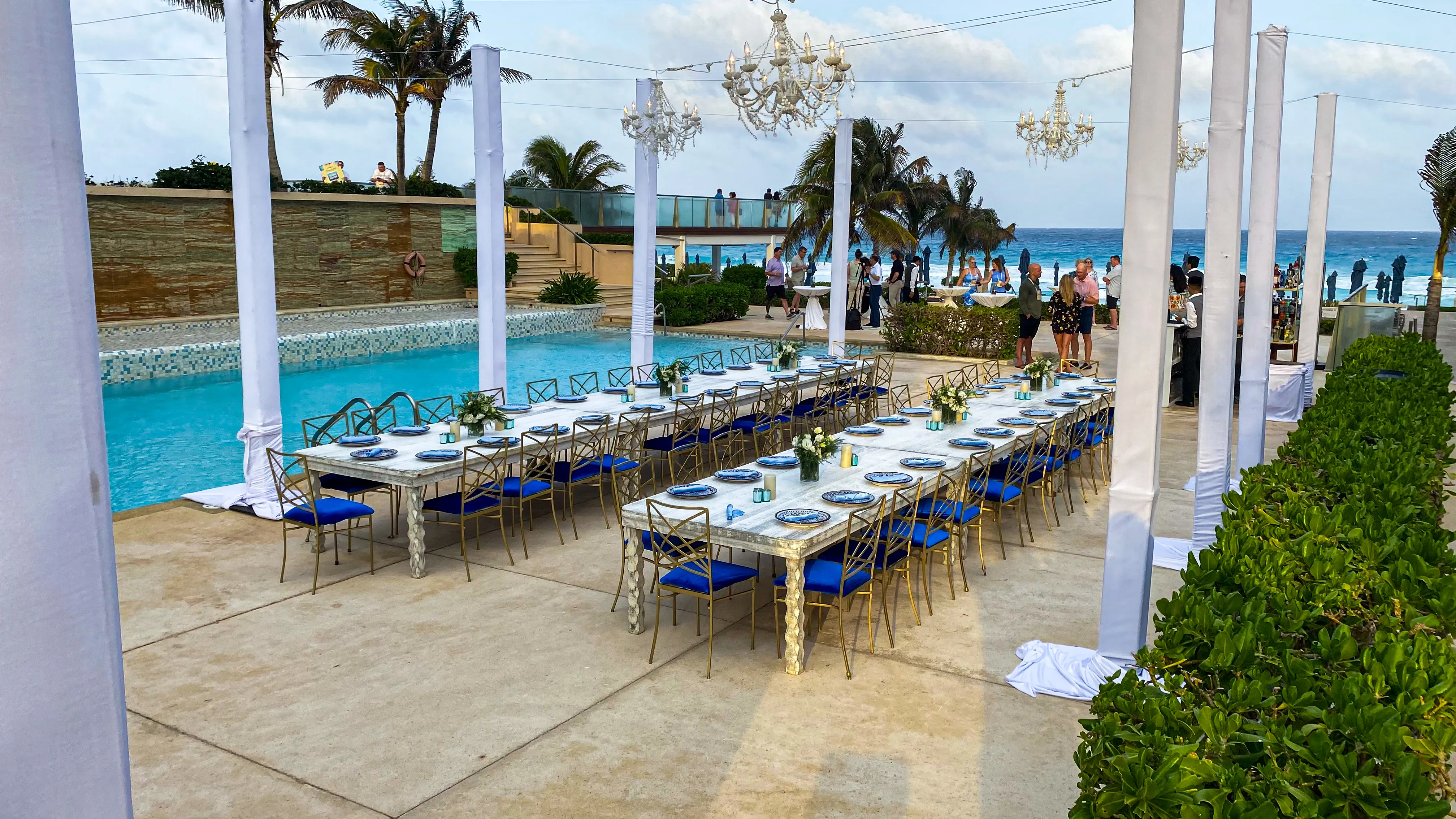 A long poolside dinner table set with blue and gold chairs beneath white chandeliers at dusk.