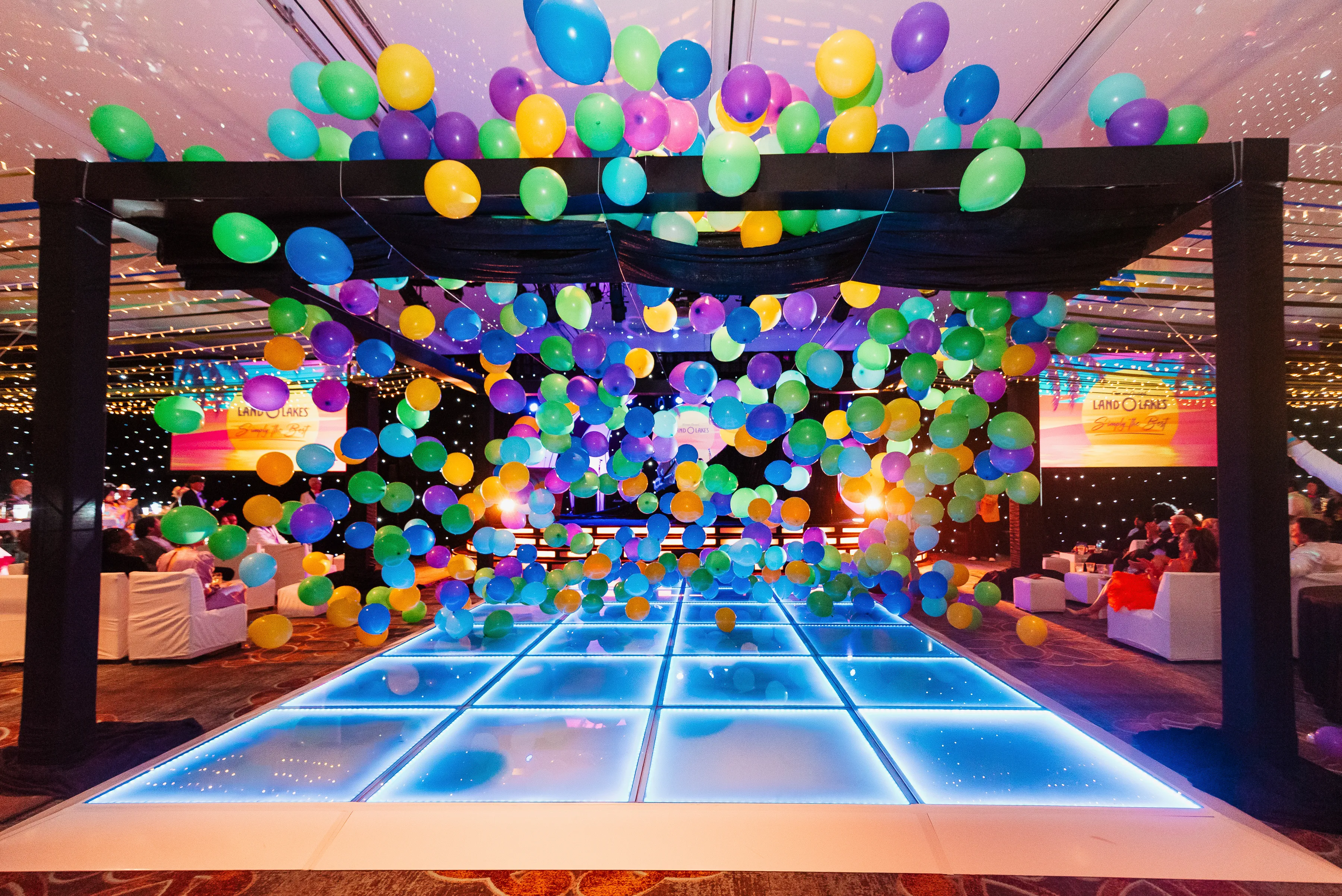 A ballroom dance floor covered in colorful balloons falling from the ceiling above dancing guests.