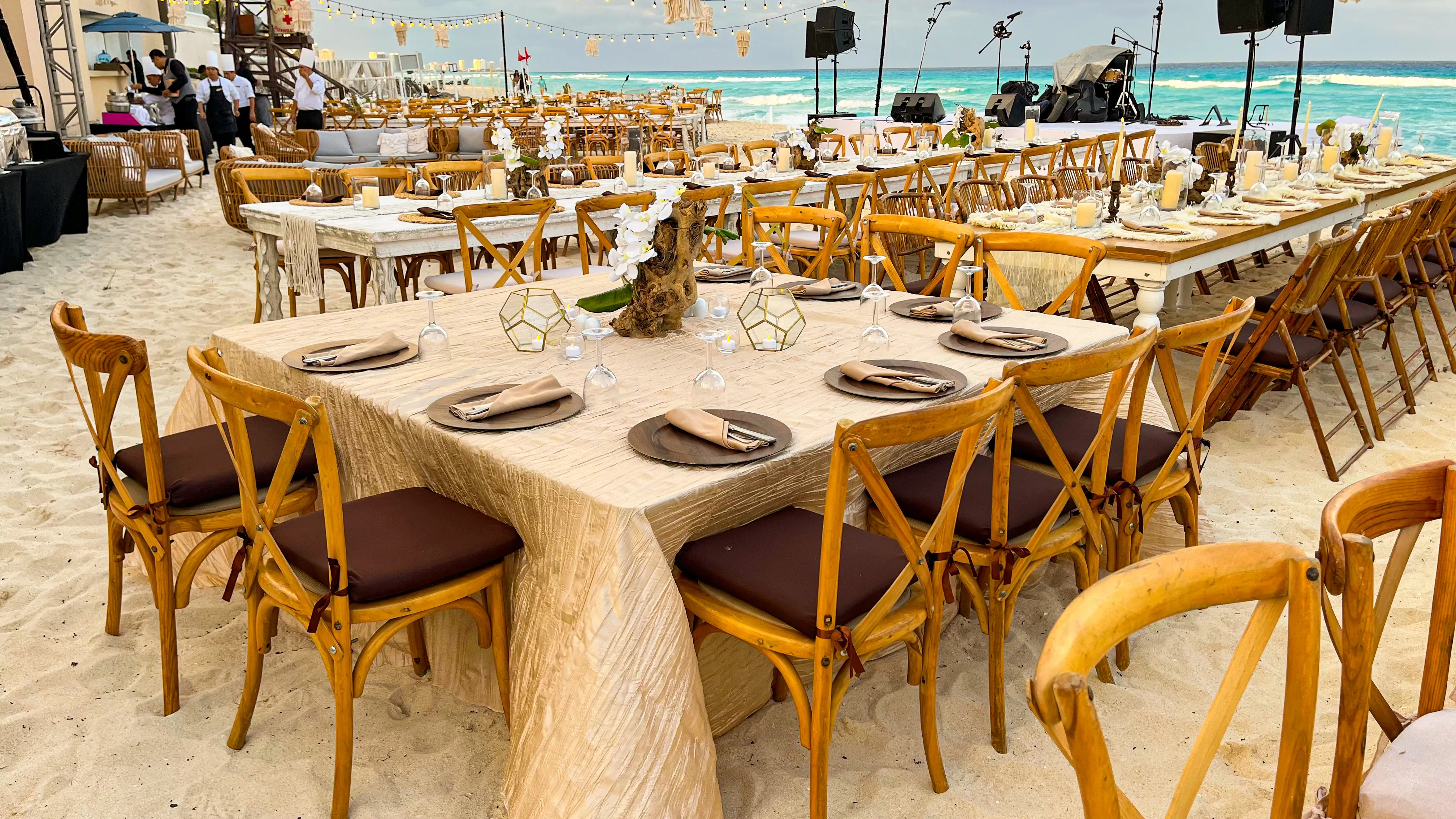 A square dining table set on the beach with wood chairs, candles, and the ocean behind it.