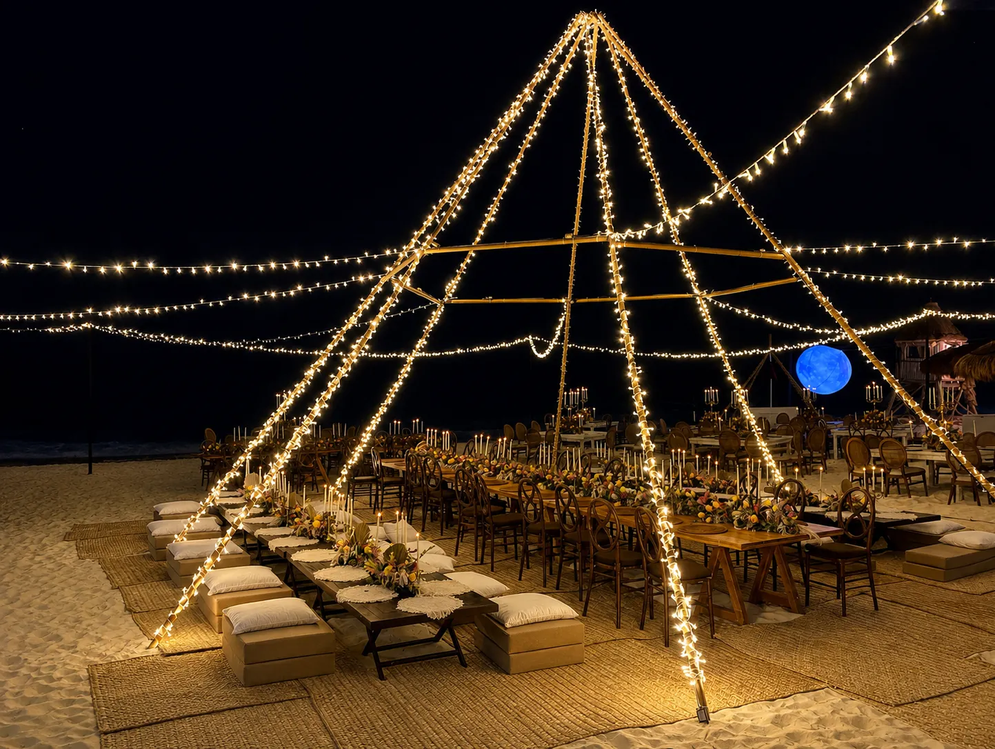 A large teepee lit at night over long low tables with cushions set on woven petate mats on the sand.