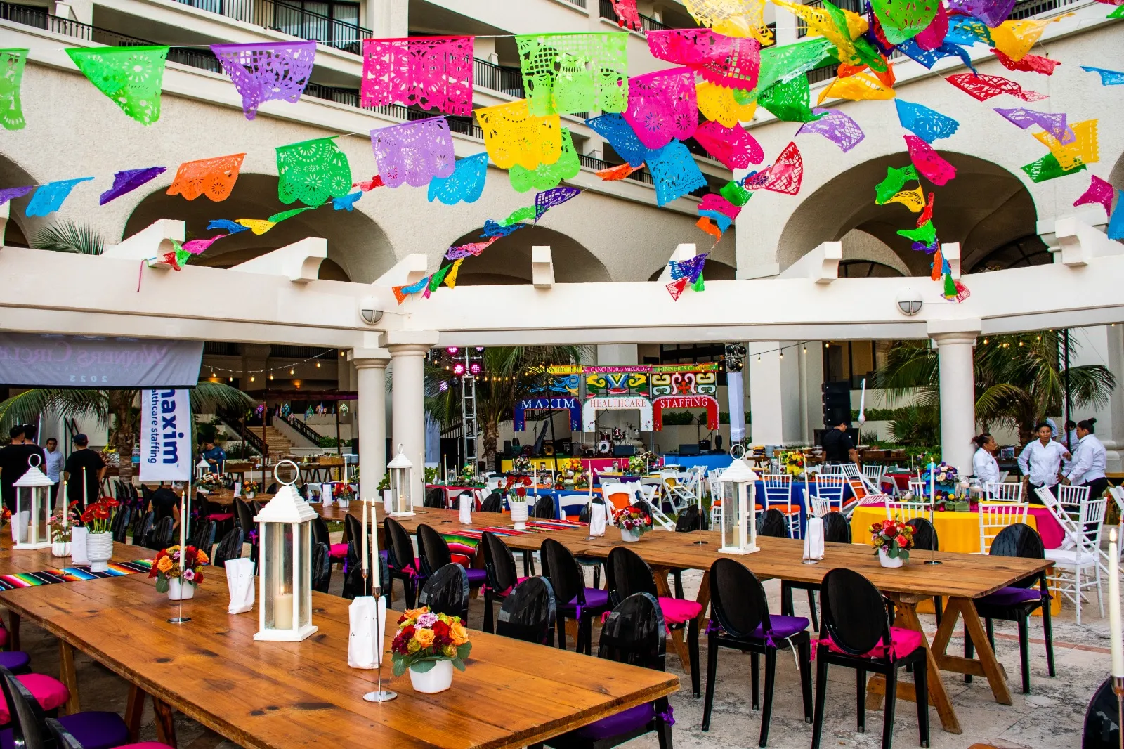 An outdoor resort walkway strung with colorful papel picado banners above rows of wooden dinner tables.