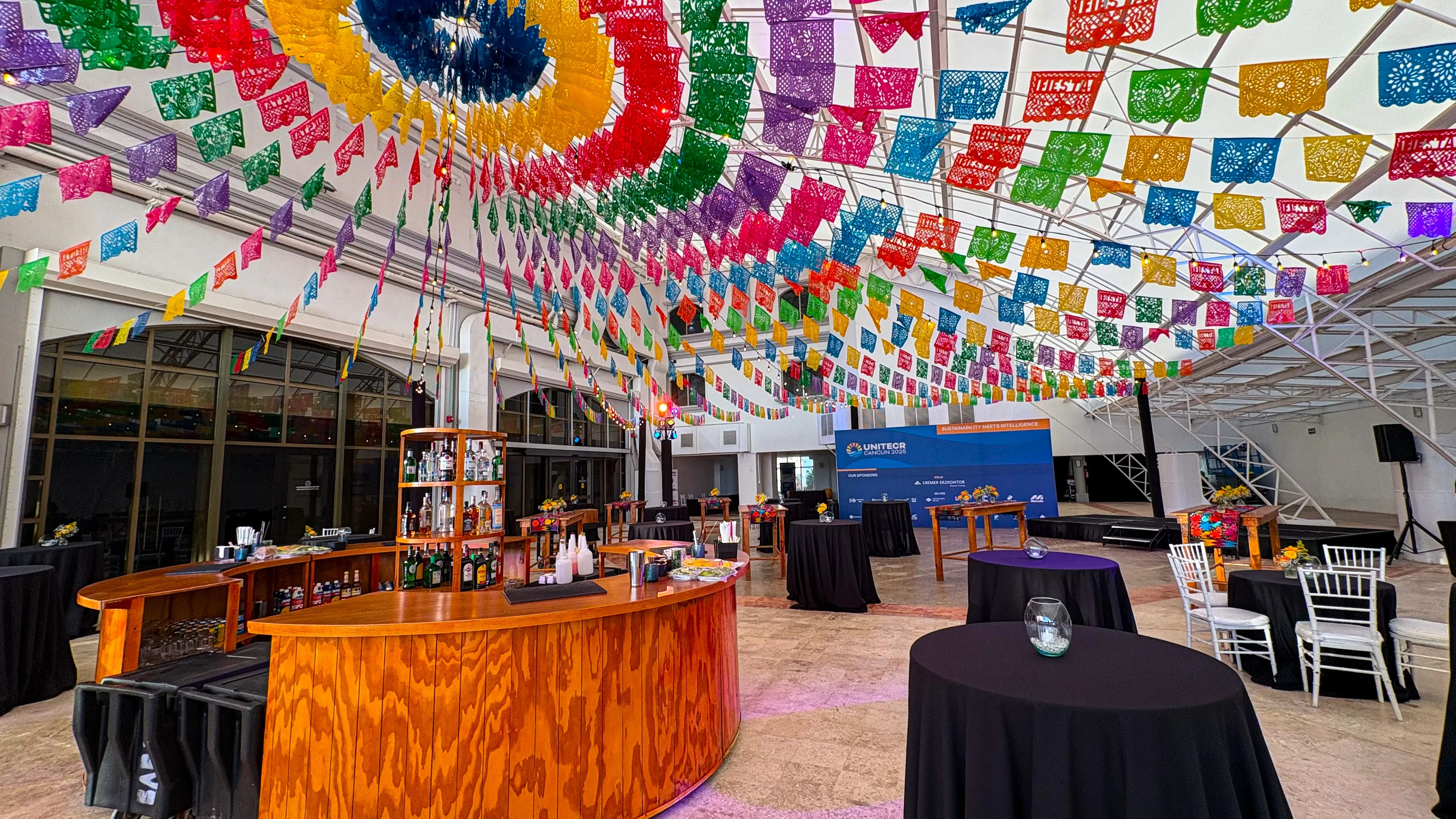 A covered terrace strung with a dense radiating canopy of papel picado above a round wooden bar and cocktail tables.