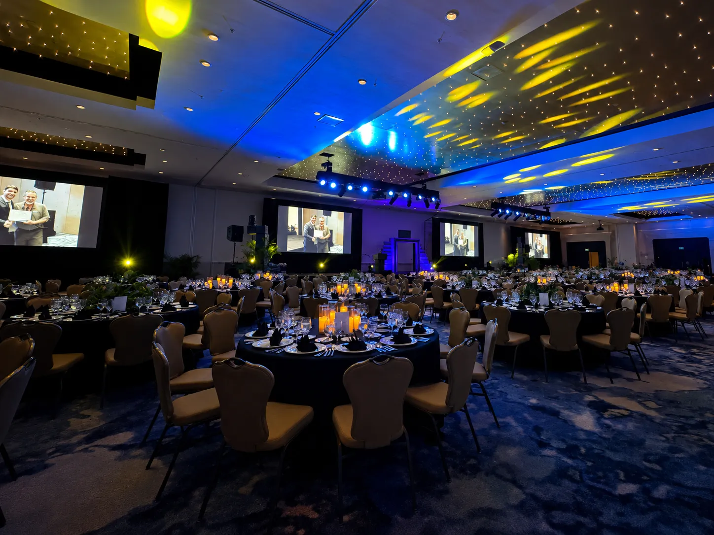 A ballroom set with round tables and chairs beneath blue lighting and a starlit ceiling for an evening dinner.