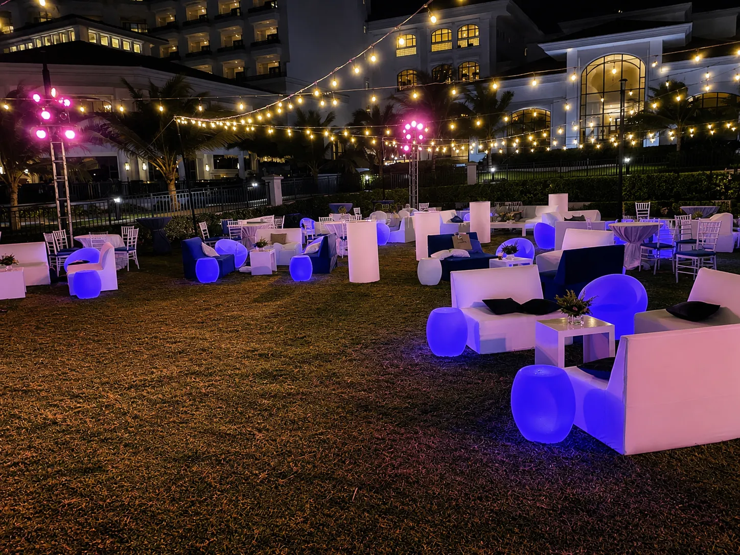 An empty garden lounge lit blue at night, with white cocktail tables and string lights strung through the space.
