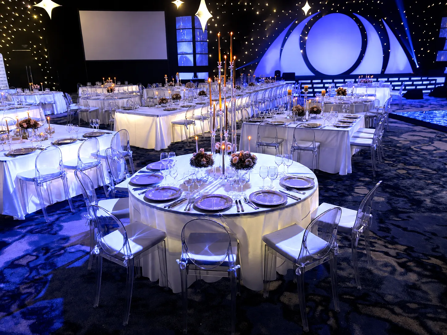 Long banquet tables with white linens and glowing centerpieces sit beneath a black ceiling scattered with hanging stars.