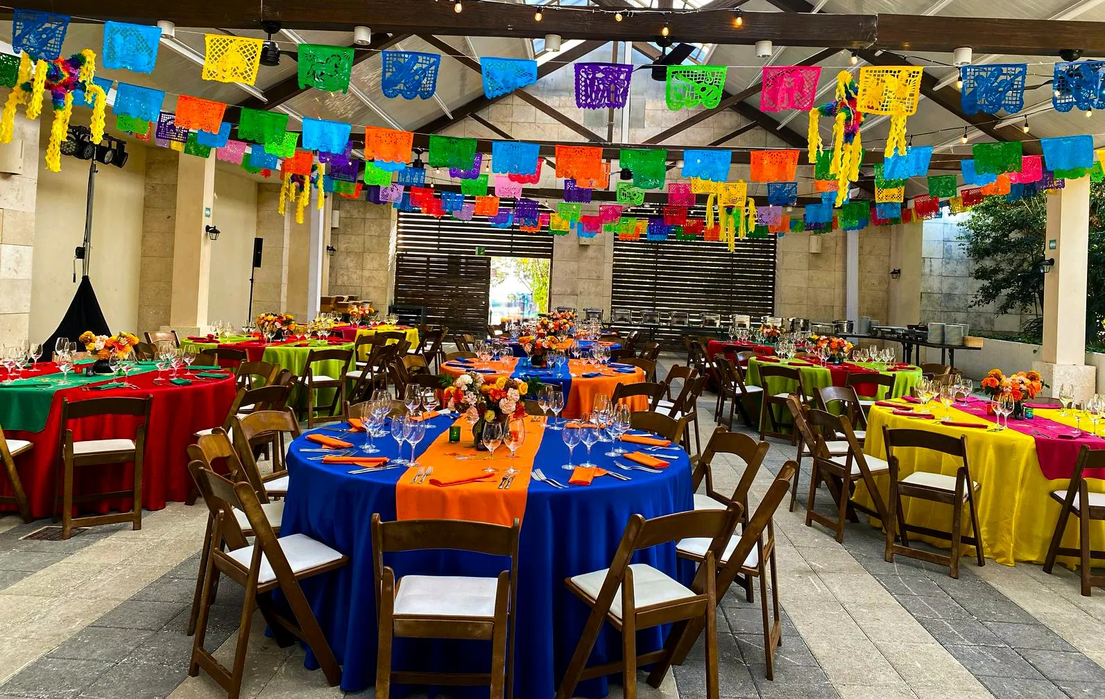 Round tables in bright linens with contrasting runners and floral centerpieces beneath papel picado on a covered terrace.