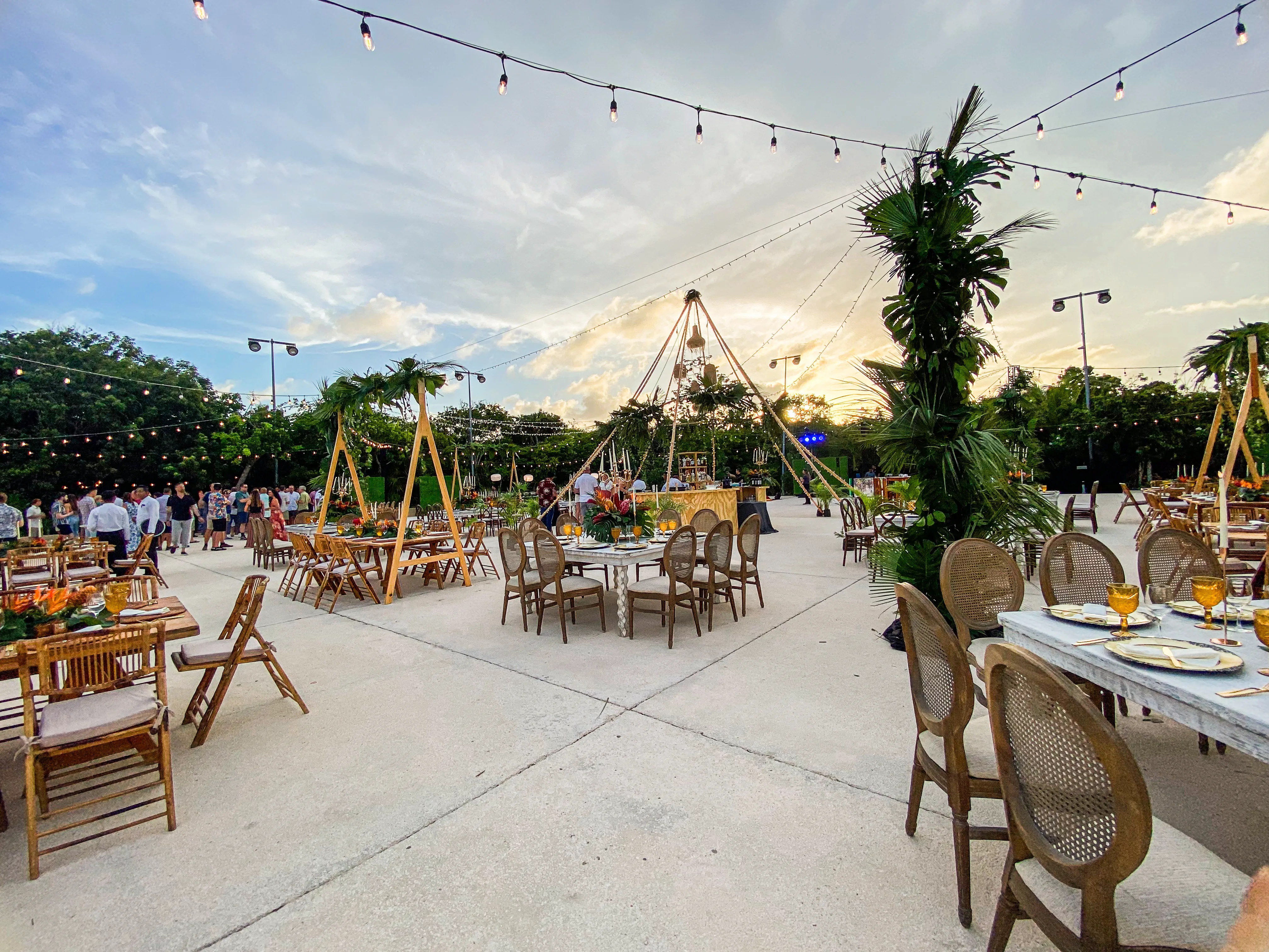 Tropical dinner setup on a paved terrace with mixed wooden tables, foliage-wrapped lighting poles, and a tall timber canopy over the bar.