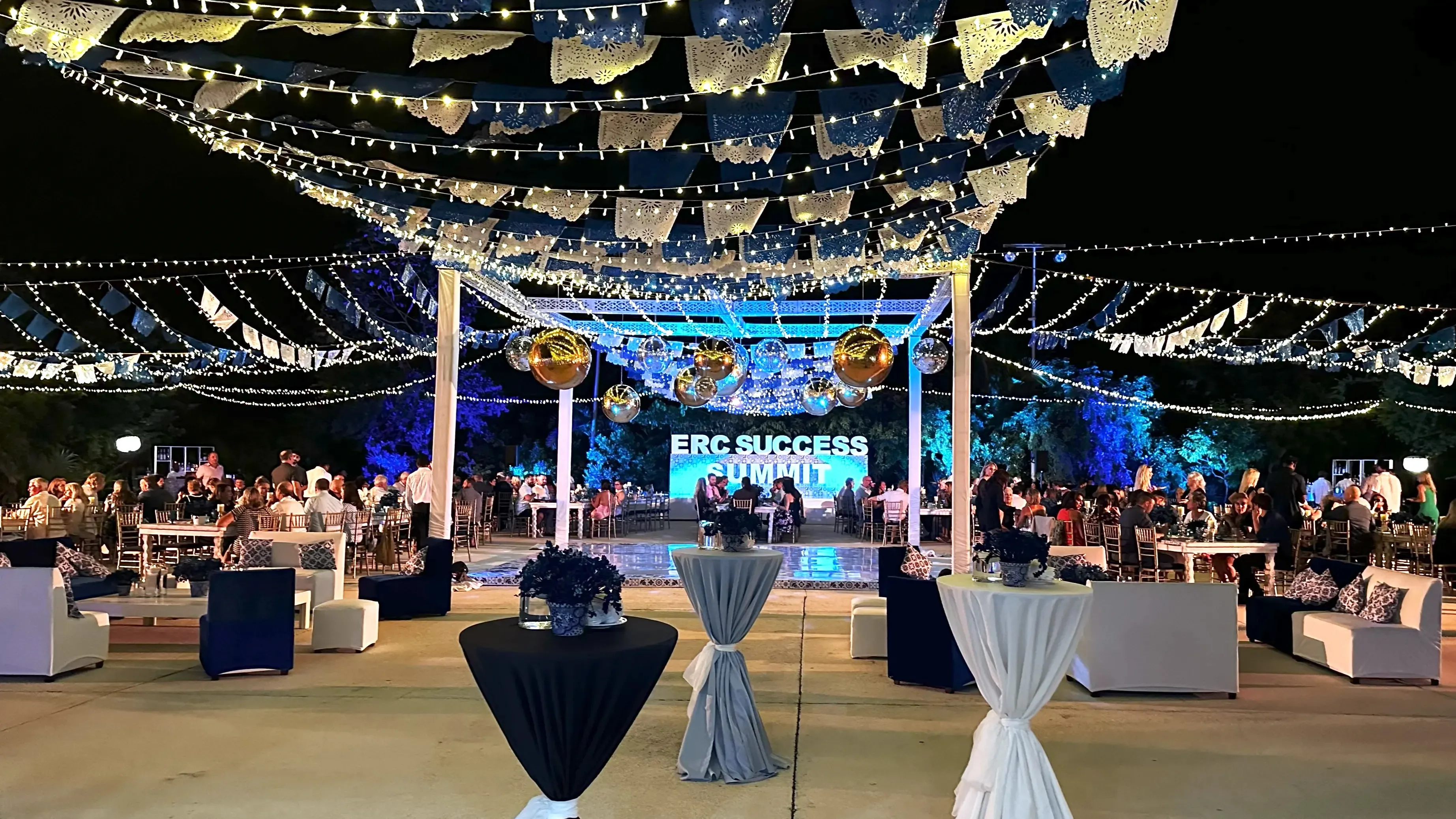Nighttime recognition dinner setup with blue and white papel picado, hanging gold spheres, a dance floor, and an illuminated stage.