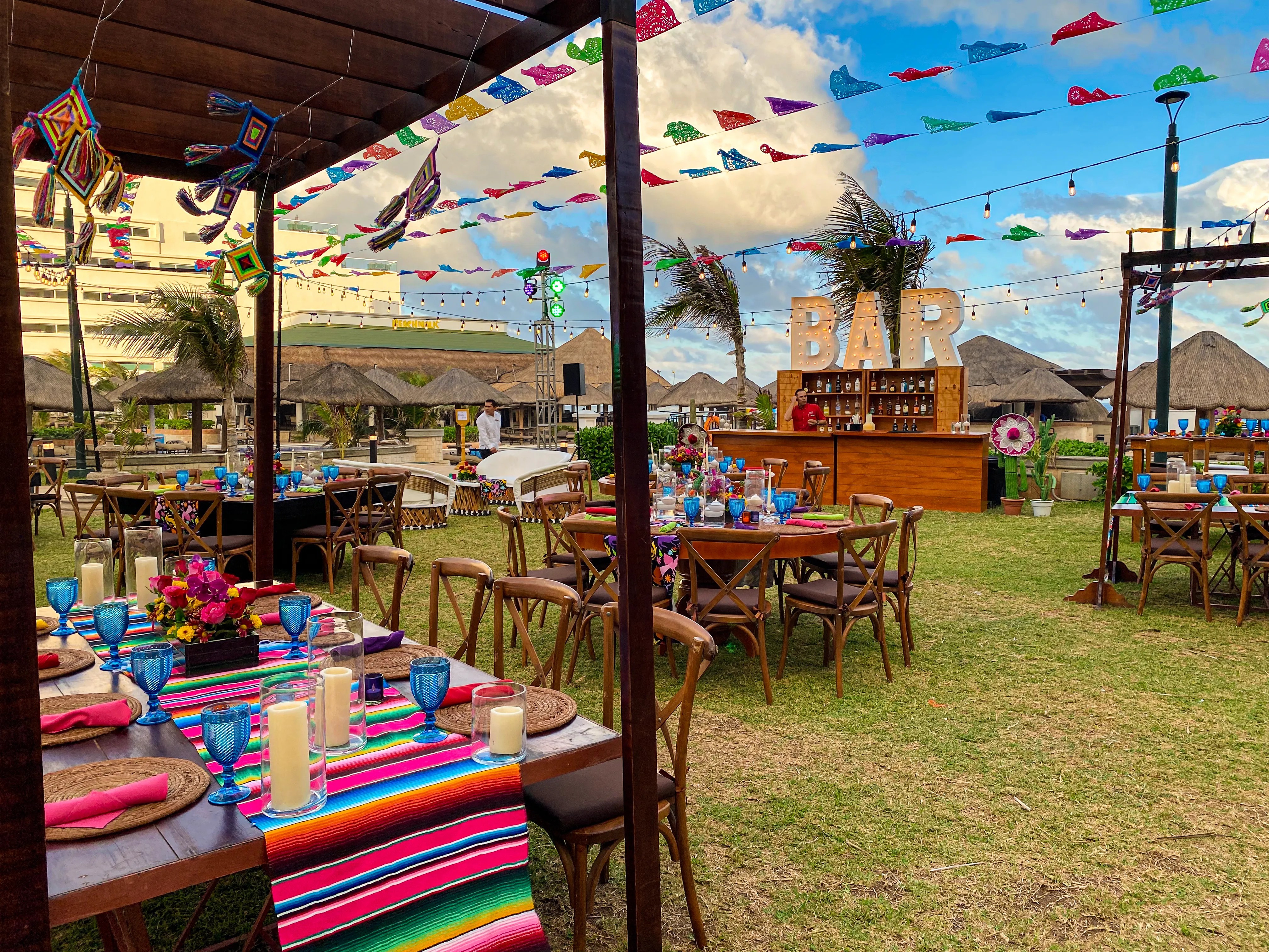 Serape-covered dining tables beneath wooden pergolas and colorful flags, with illuminated bar lettering in the garden beyond.