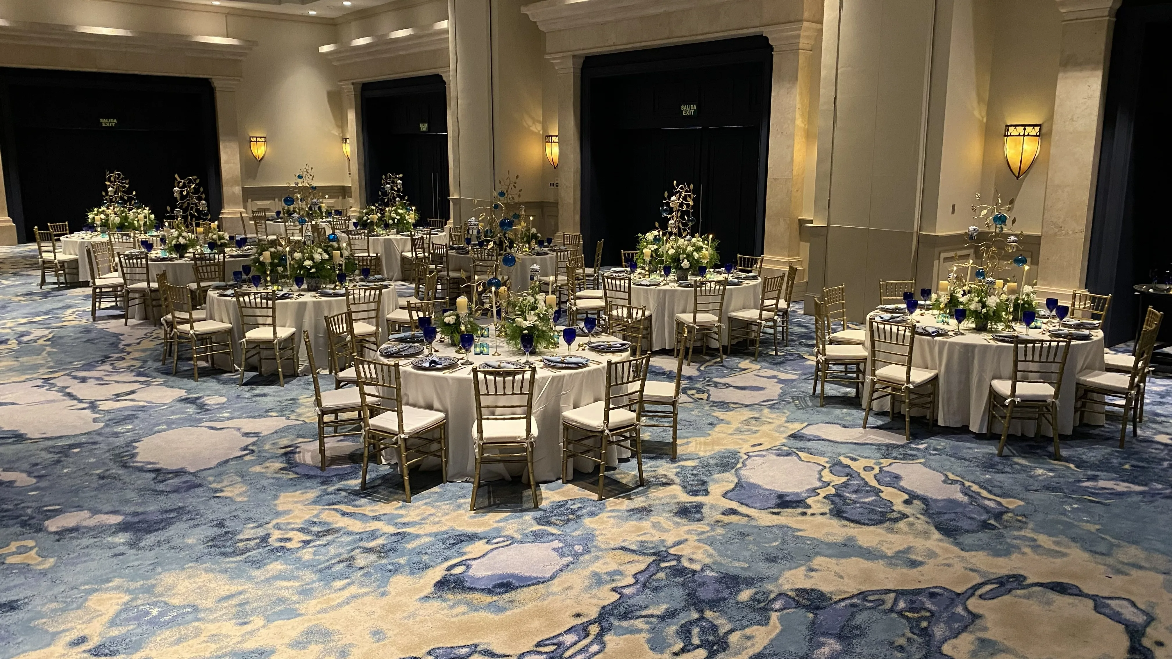 Round lunch tables in white linens with gold chairs, blue glassware, and Talavera-inspired table decor in Tulum Ballroom.