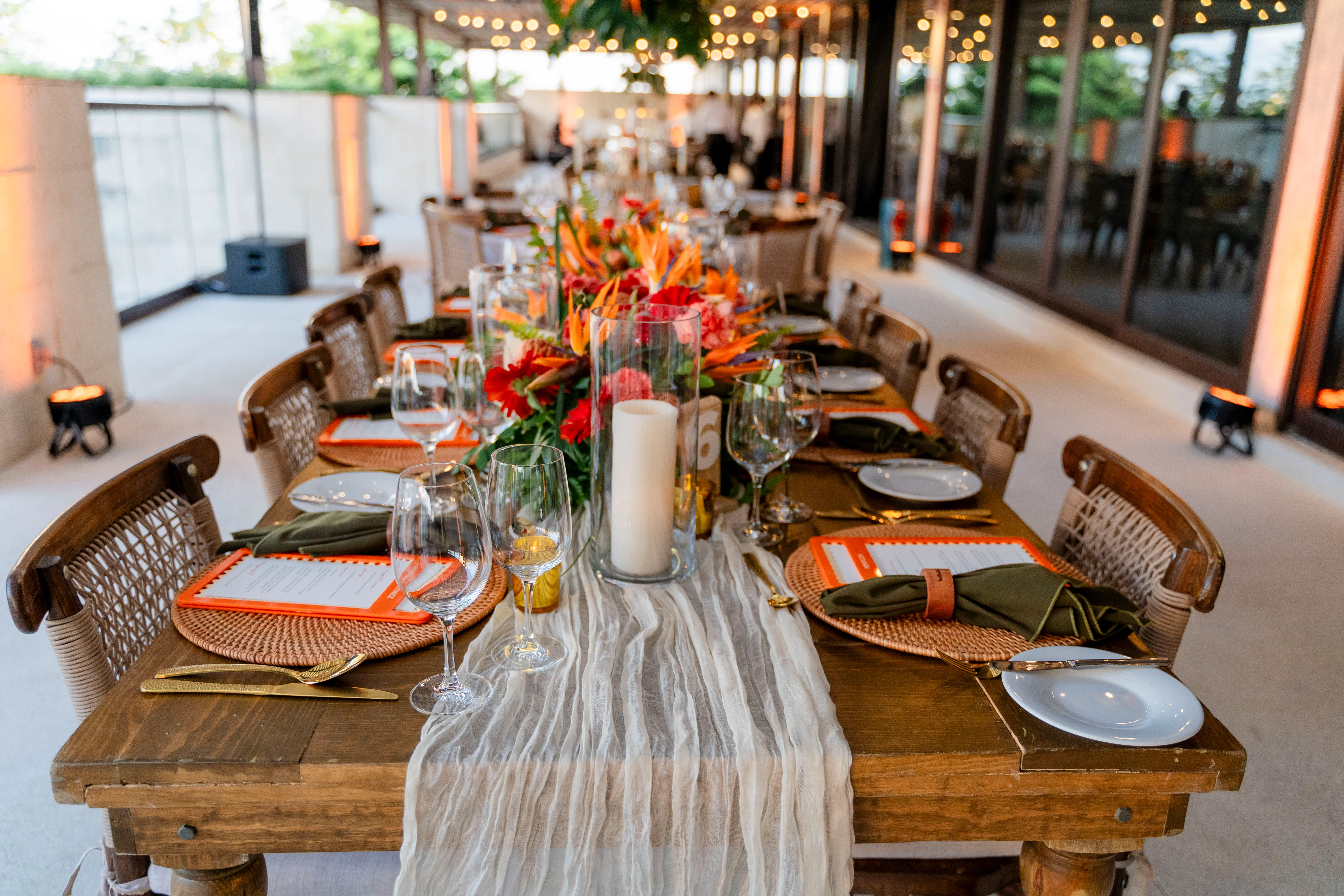 A long wooden dinner table with a gauze runner, woven chargers, orange menus, and tropical flowers on a covered terrace.