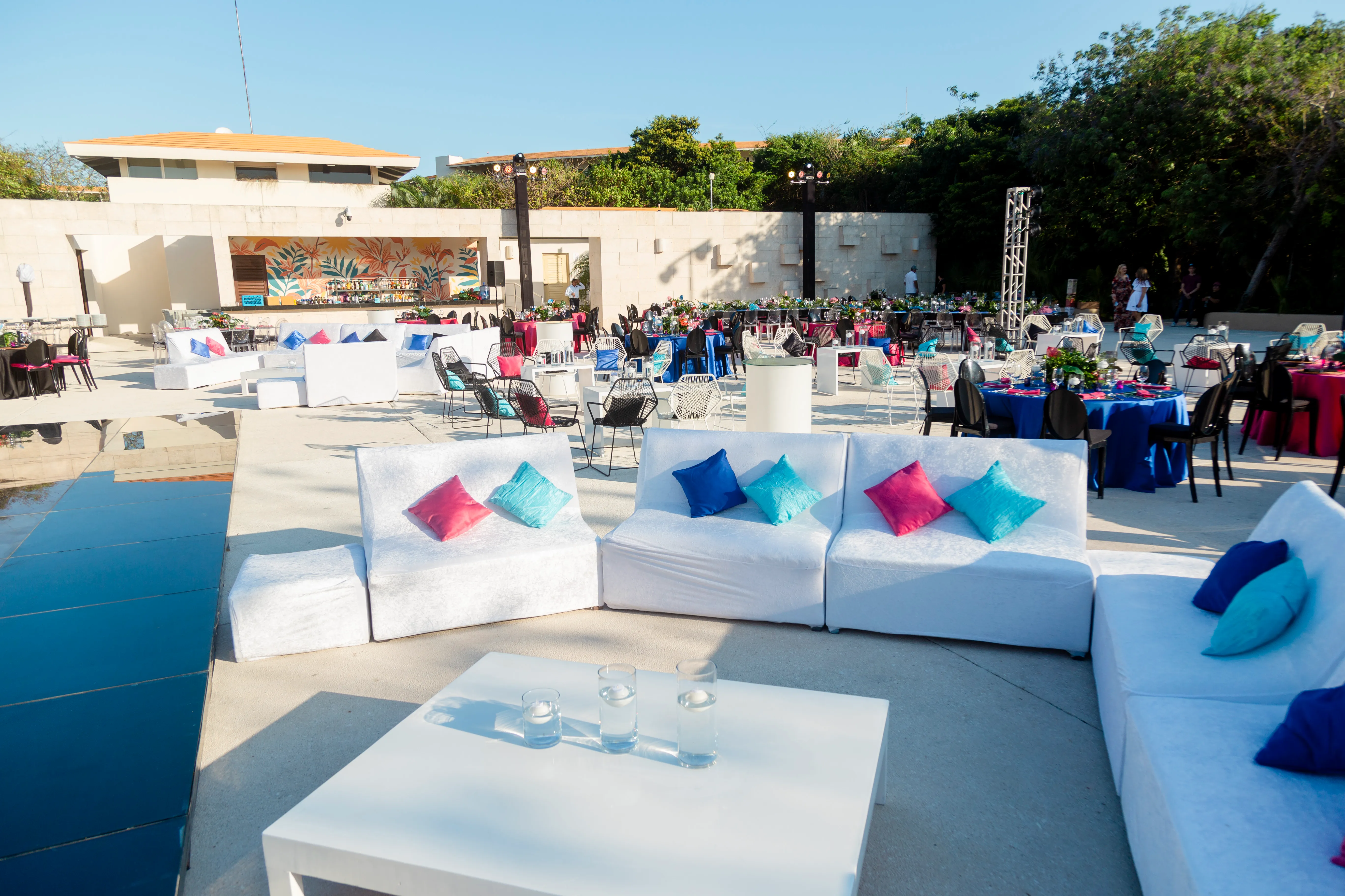 White lounge sofas with blue and pink cushions beside black dining tables during an outdoor welcome event setup.