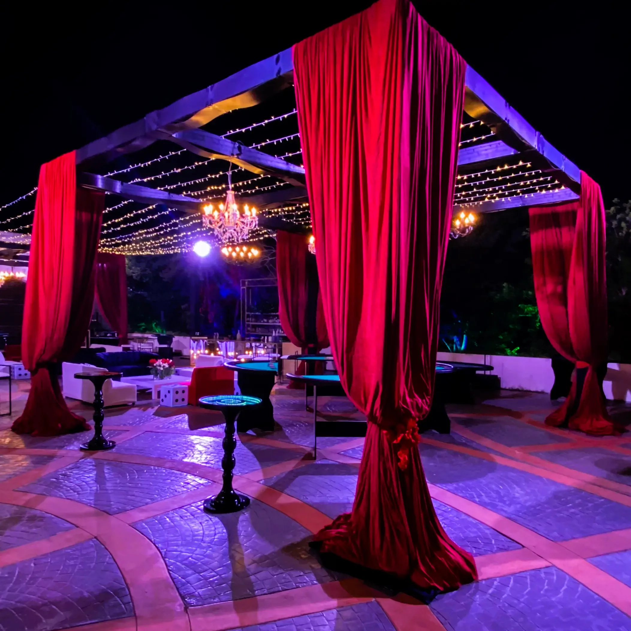 Red velvet curtains frame a patio gaming area beneath chandeliers and strings of fairy lights at night.