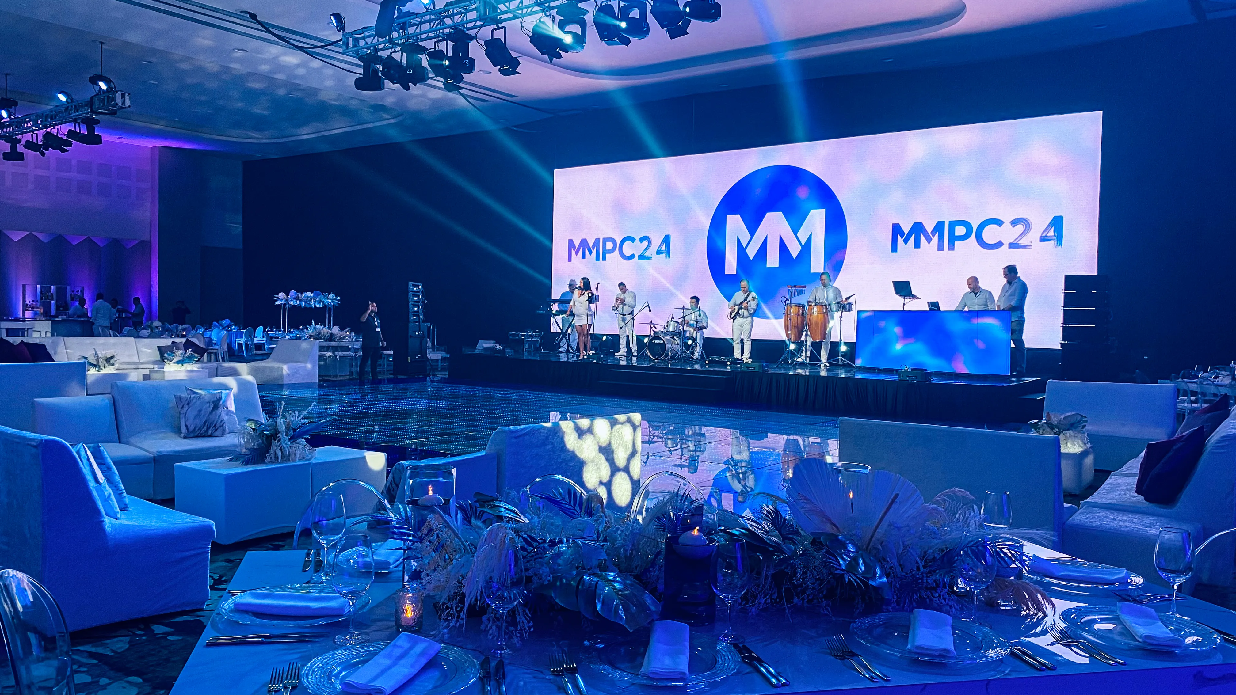 A ballroom dinner setting with pale tropical foliage, glass chargers, and white seating under blue lighting, facing an illuminated stage.