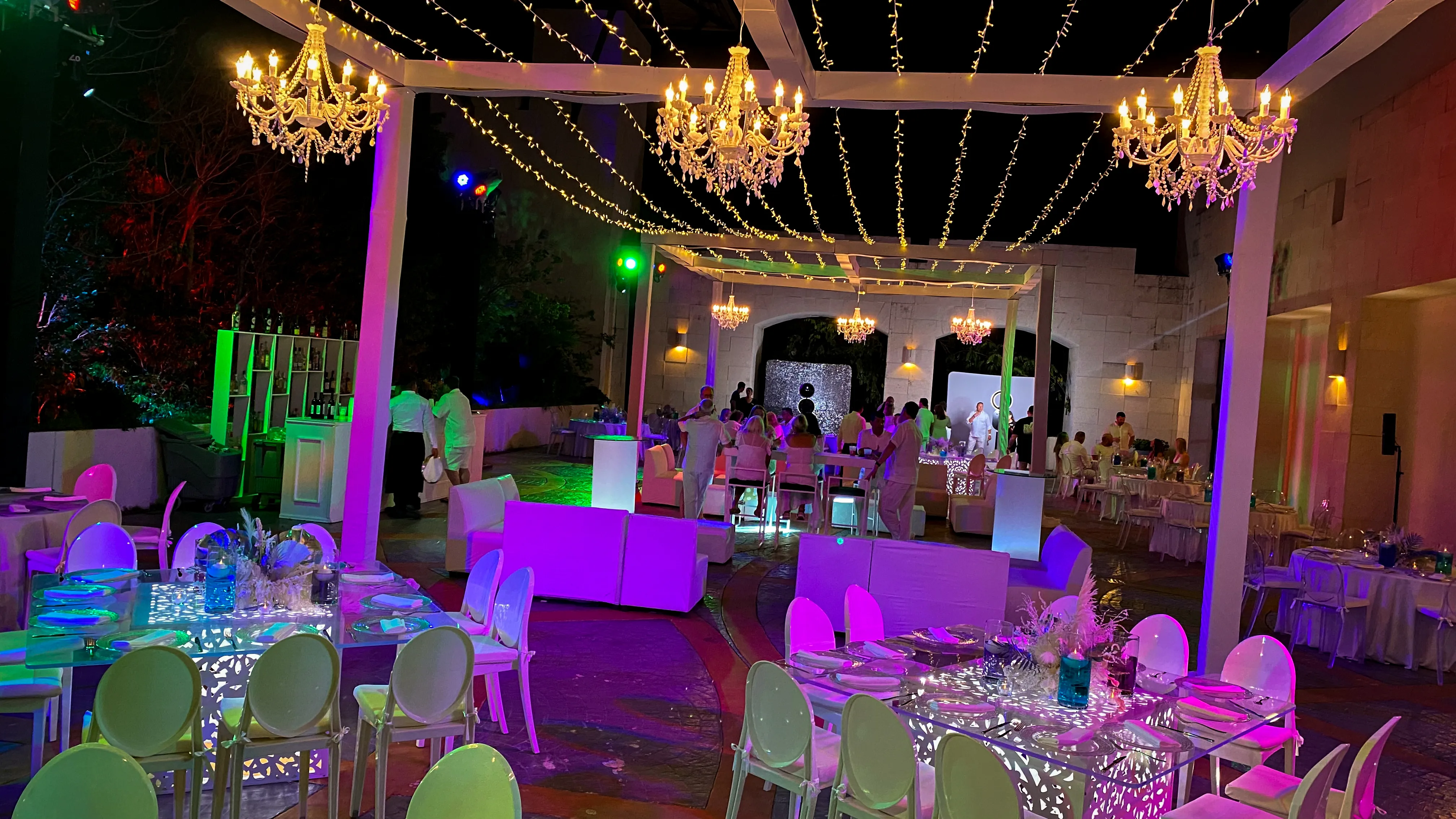 A patio dinner setting beneath white-clad structures and chandeliers, with fairy lights and colorful lighting at night.