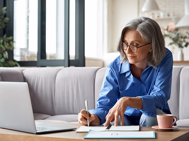 Mature woman with glasses sitting on sofa, writing on paper with laptop and coffee cup on table.
